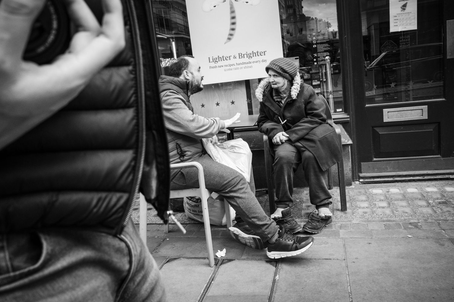Black-and-white street photo of two people in conversation outside a London café, capturing candid urban life and human connection.