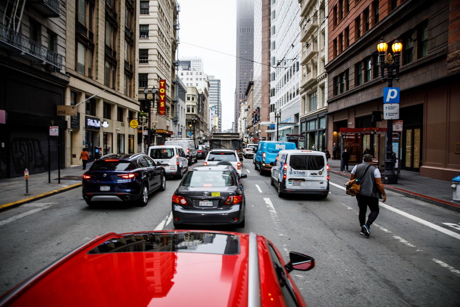 Downtown San Francisco street scene with traffic, a red convertible, foggy skyscrapers, and a pedestrian crossing the road.