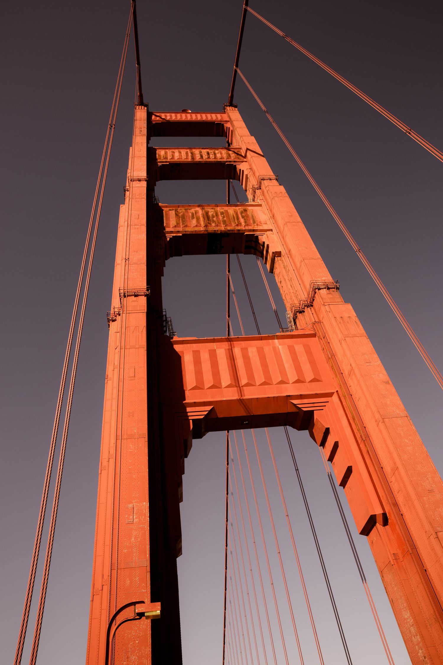 Golden Gate Bridge south tower glowing in International Orange at sunset, viewed from below with suspension cables in San Francisco.