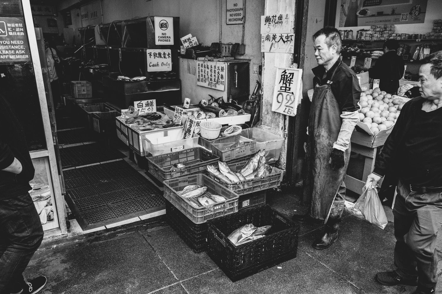 Black and white street photograph of a fishmonger outside a seafood market in San Francisco Chinatown as shoppers pass by.