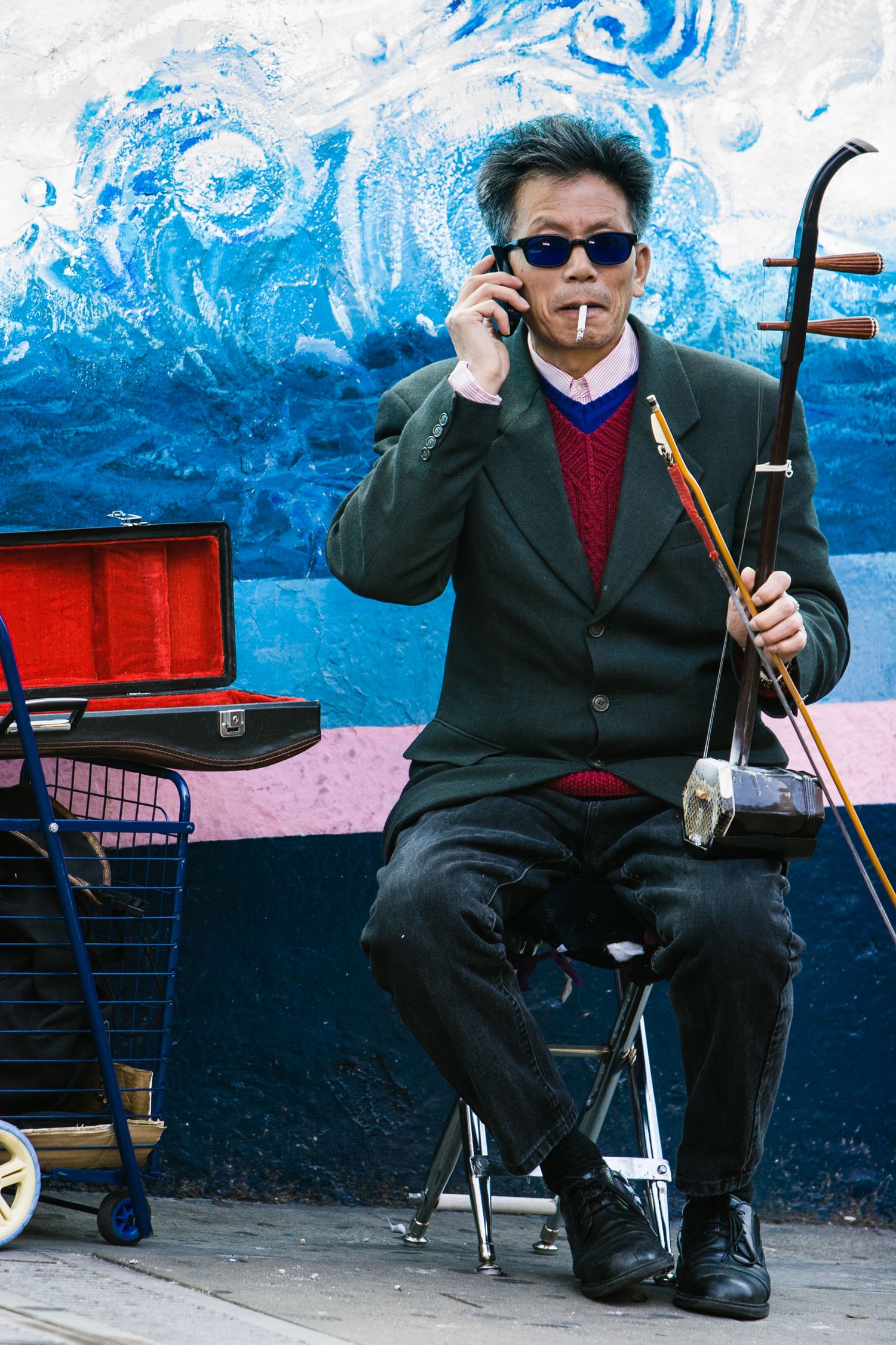 Street musician holding an erhu while talking on a cell phone during a Chinatown event in San Francisco, framed against a vibrant blue mural