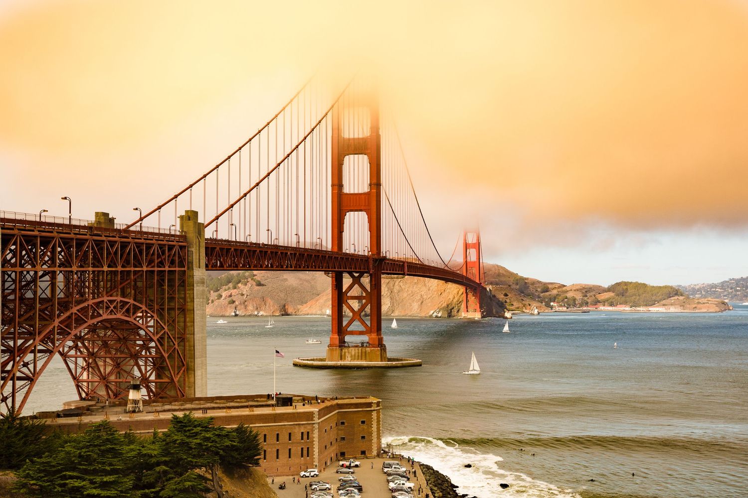 Golden Gate Bridge emerging through golden fog above San Francisco Bay, with sailboats, Fort Point, and the Marin Headlands beyond.
