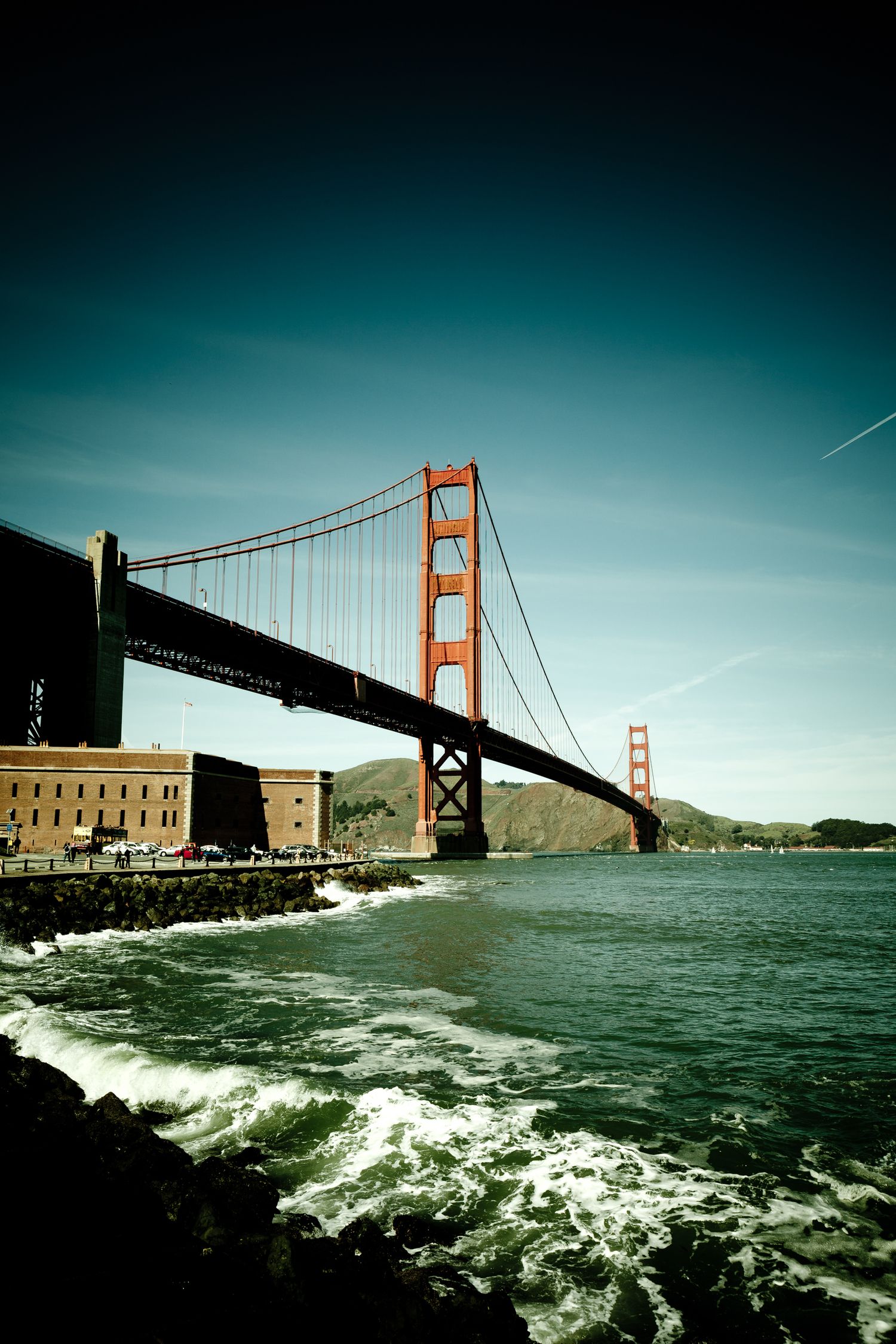 Golden Gate Bridge rising above waves at Fort Point, with San Francisco Bay and Marin Headlands under a clear blue sky.
