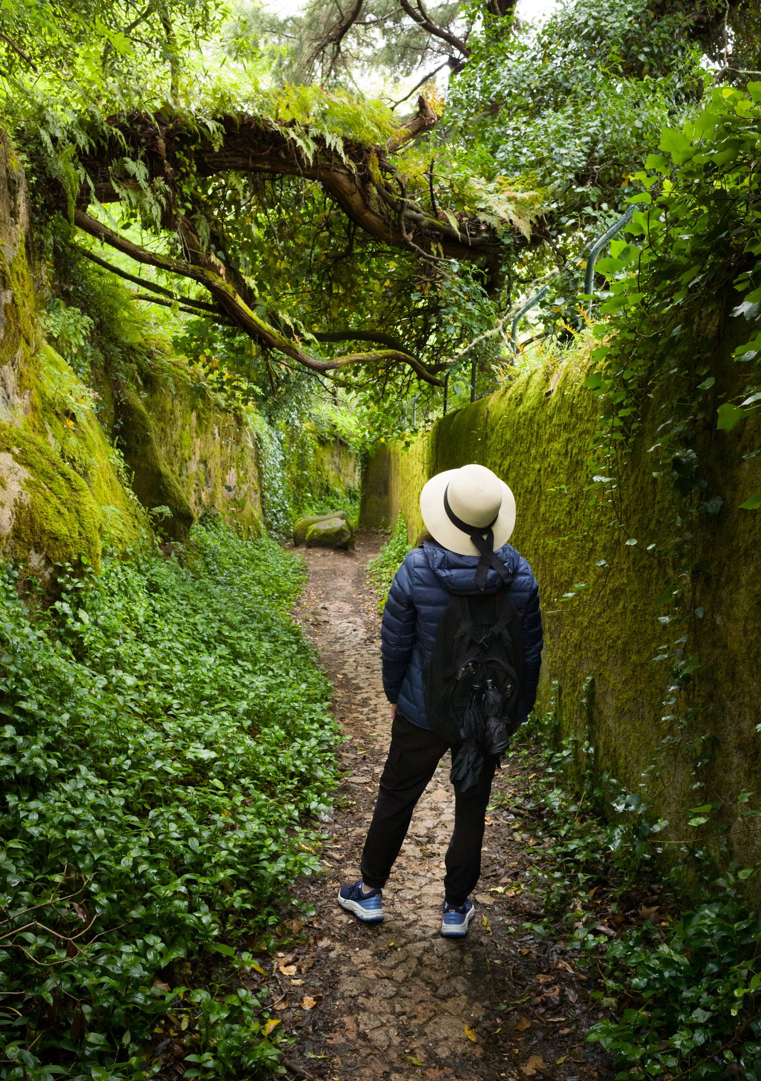 Woman in a white hat walking a moss-covered path at Quinta da Regaleira, Sintra, Portugal, in FotoMunki's Hat Series.