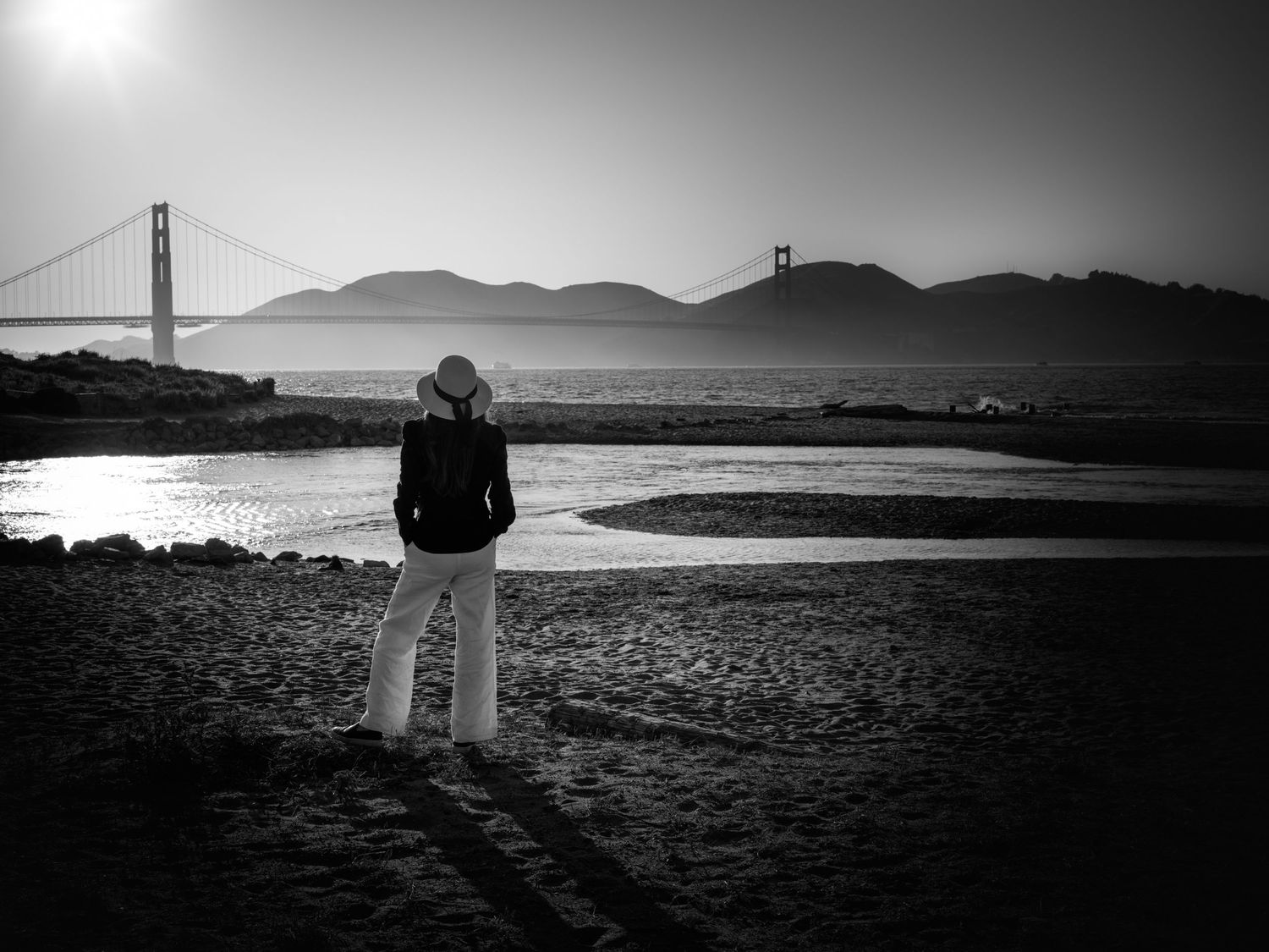 Woman in white hat overlooking the Golden Gate Bridge from Crissy Field, San Francisco, in a dramatic black and white landscape.