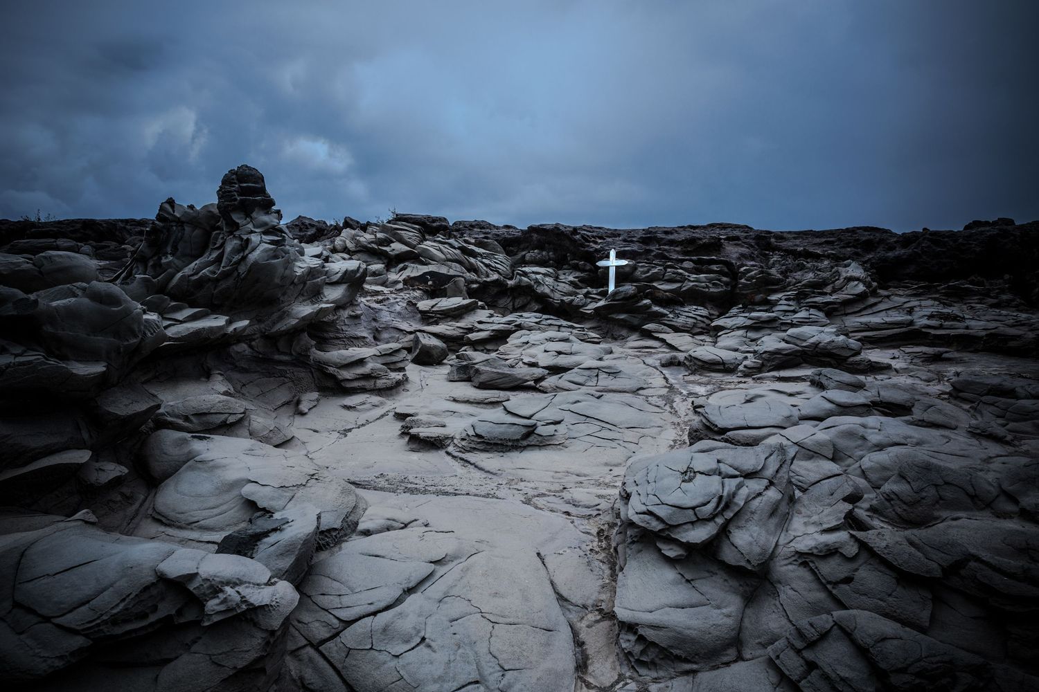 White cross on Maui volcanic lava rock beneath dramatic storm clouds in a moody fine art landscape.