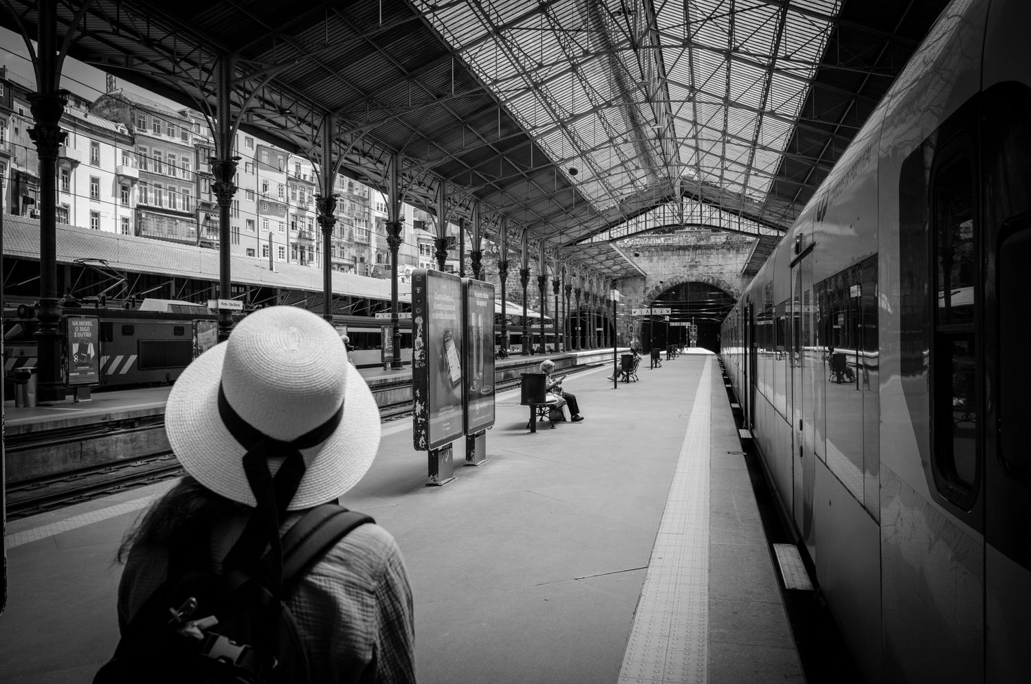 Woman in white sun hat waits beside a train at Porto, Portugal station, framed by dramatic architecture in black and white.
