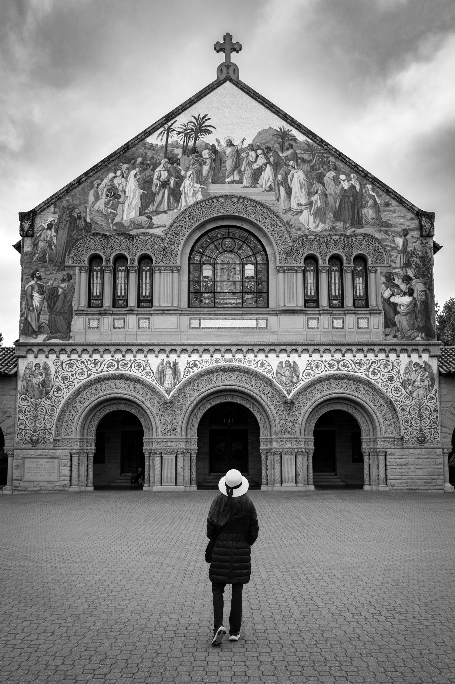 Woman in a white hat faces Stanford Memorial Church beneath dramatic skies in a black-and-white image from The Hat Series.
