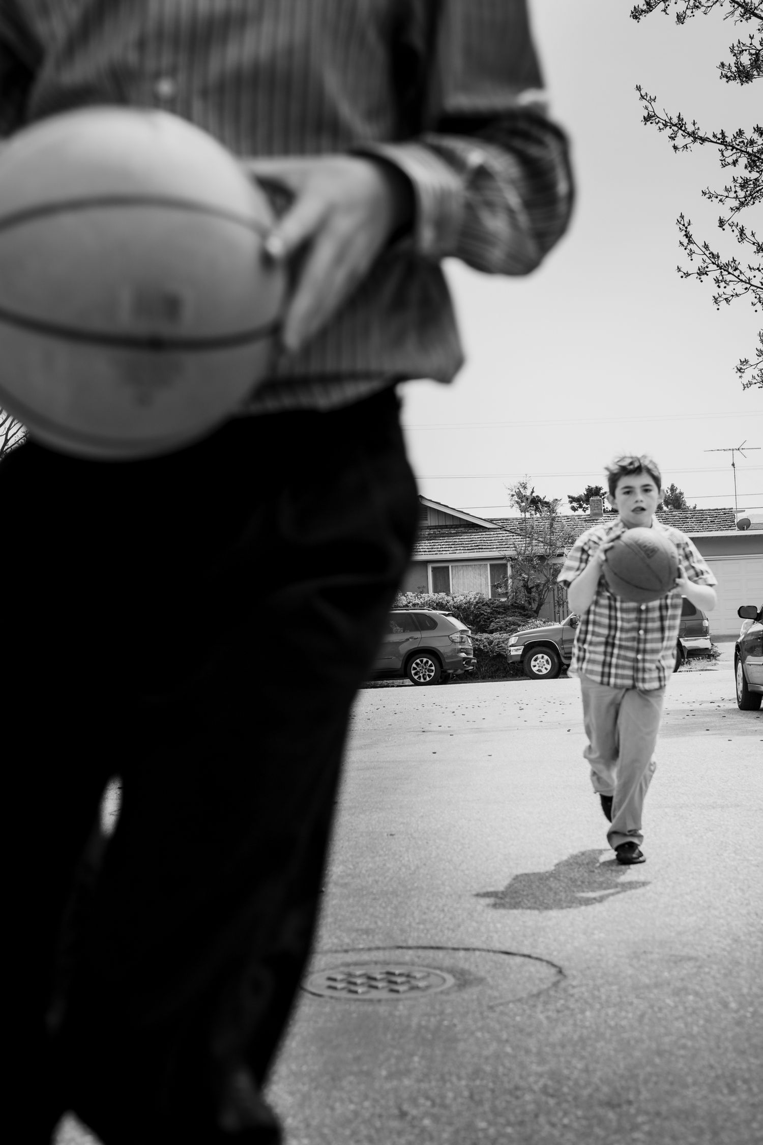 Black-and-white street photo of a child running with a basketball toward an adult, capturing play, motion, and family life in suburbia.