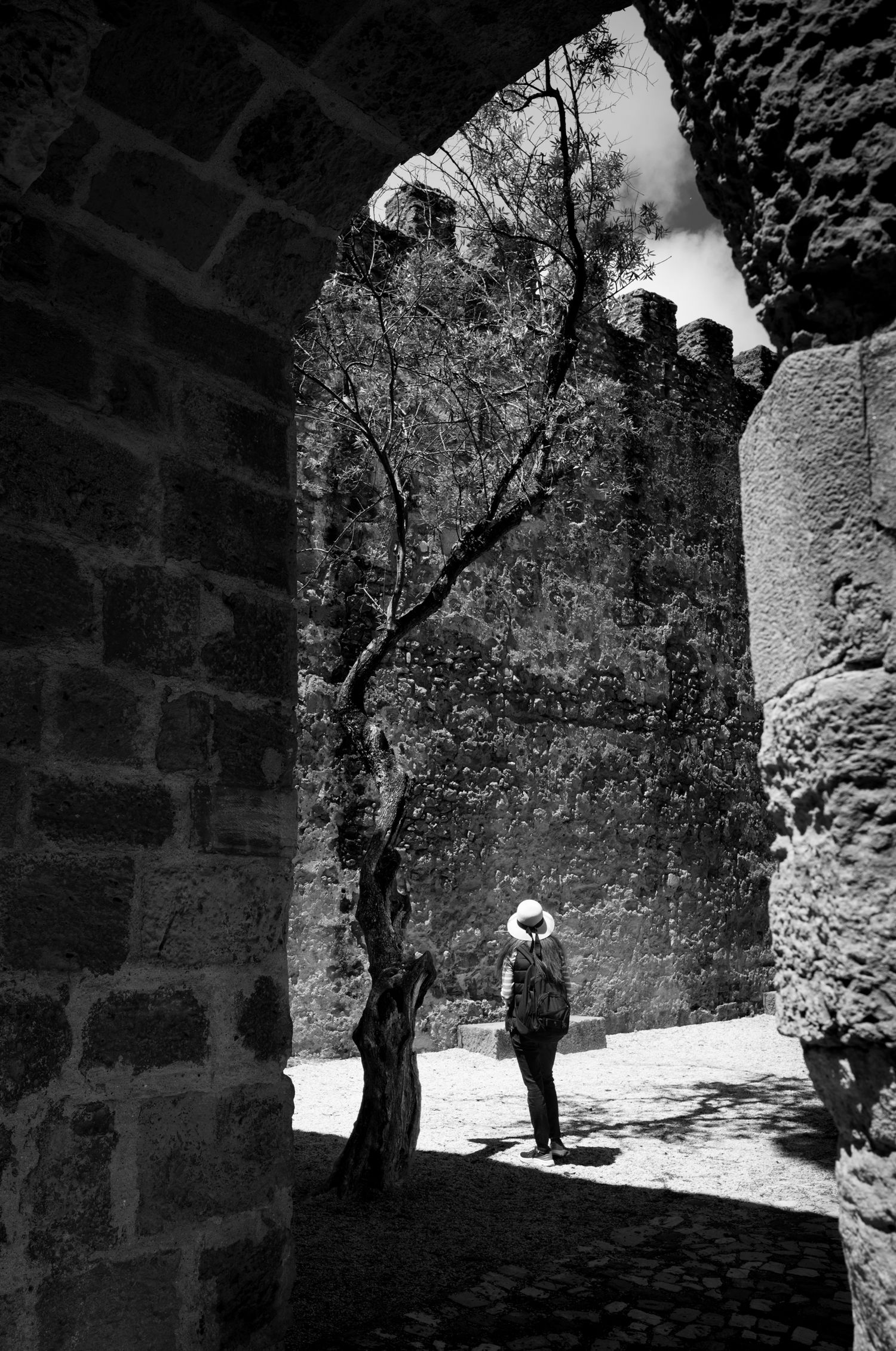 Woman in a sun hat framed by a stone arch at Castelo de São Jorge, Lisbon, Portugal, in a dramatic black-and-white scene.