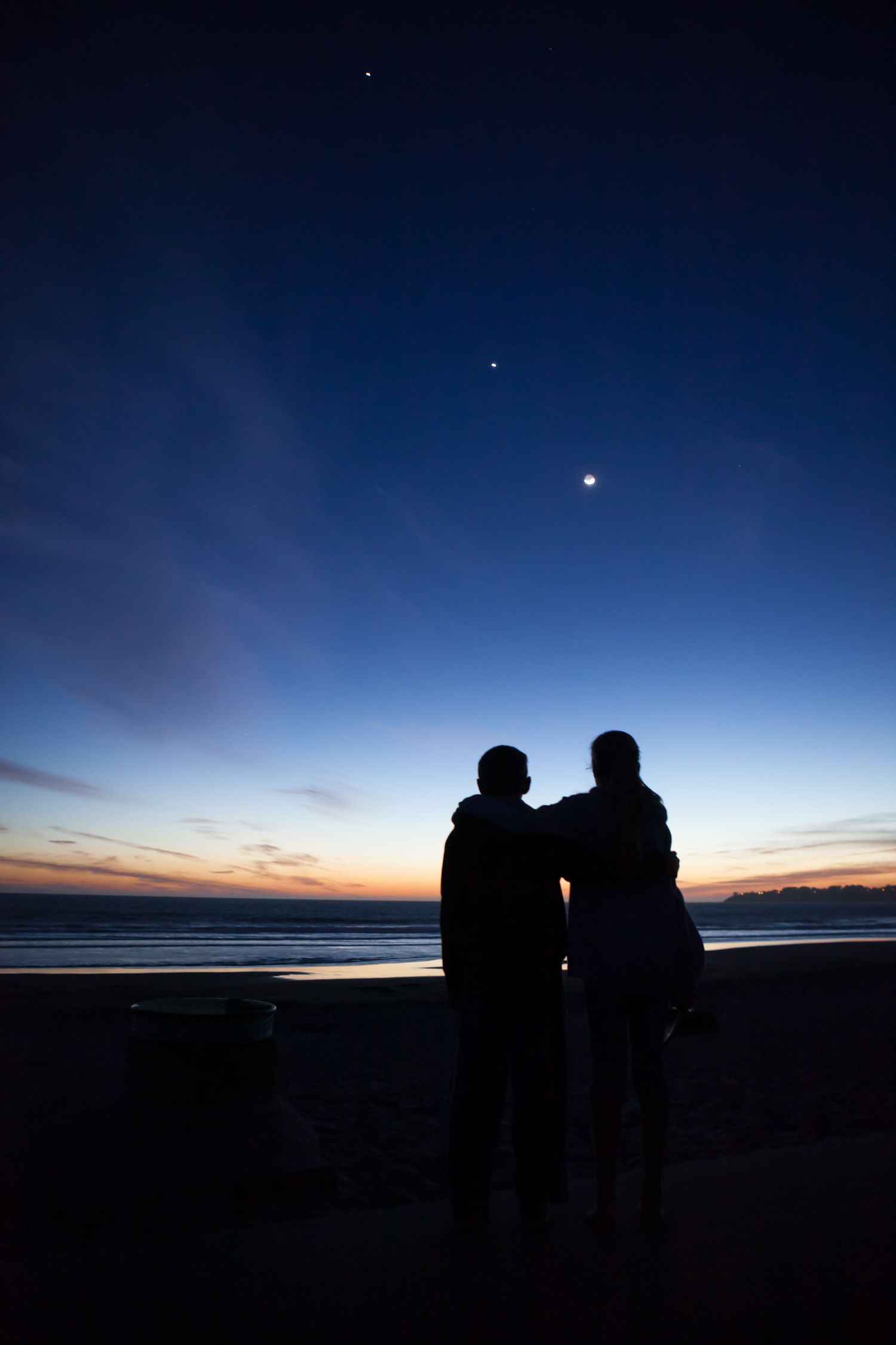 Silhouetted couple watches the crescent moon and planets over the Pacific Coast at twilight as waves meet the evening shoreline.