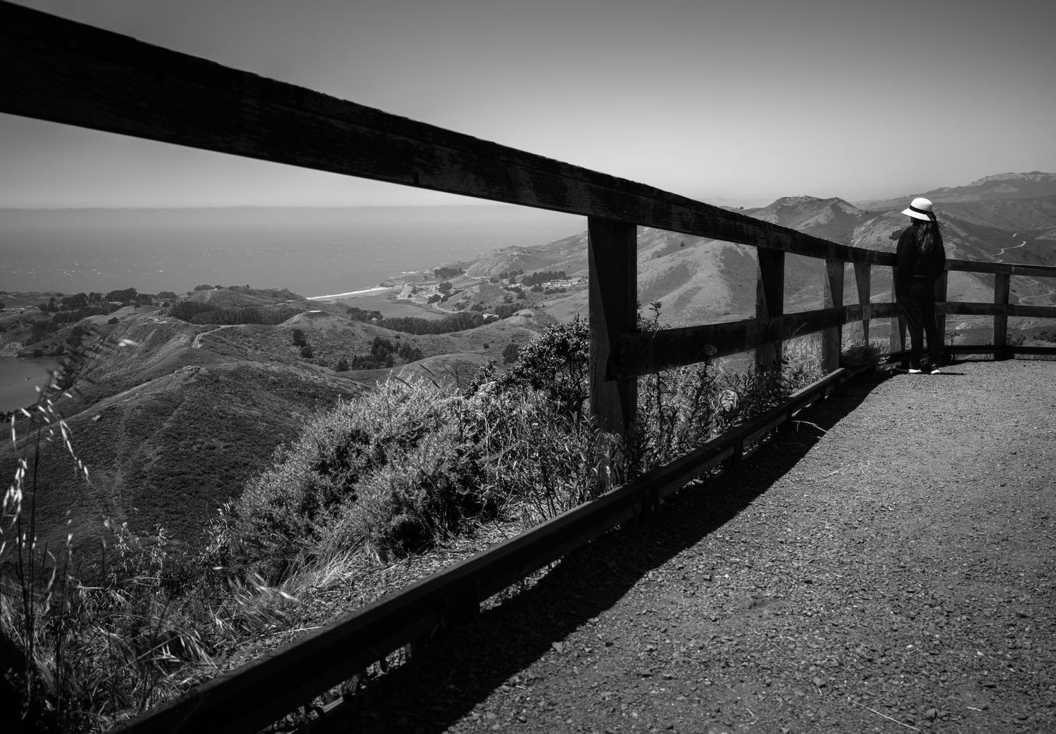 Woman in a white hat overlooks the Marin Headlands framed by the fence near San Francisco in a dramatic black-and-white coastal landscape.