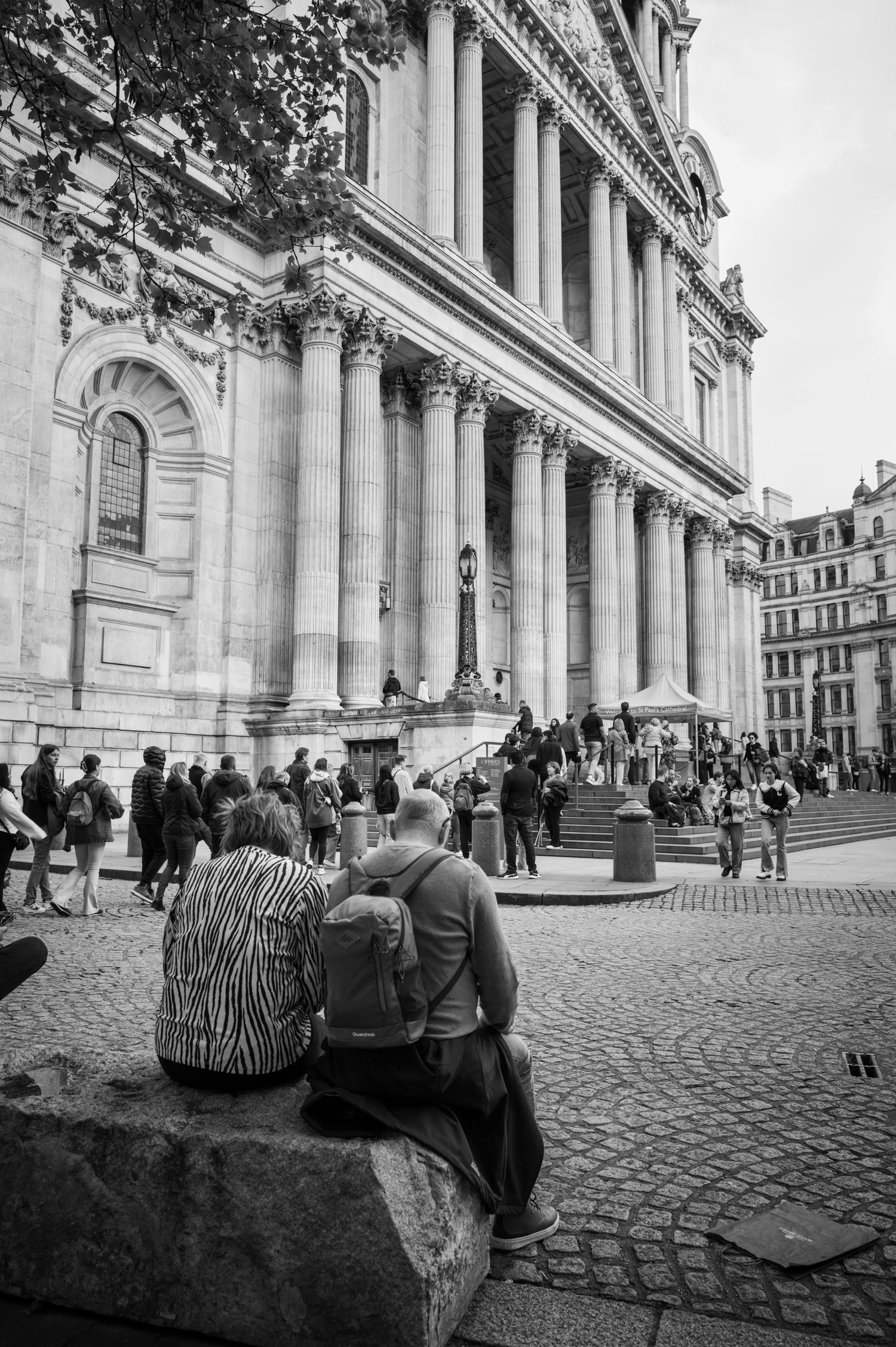 Tourists rest outside St. Paul's Cathedral in London as crowds gather beneath its towering columns in a black and white street scene.