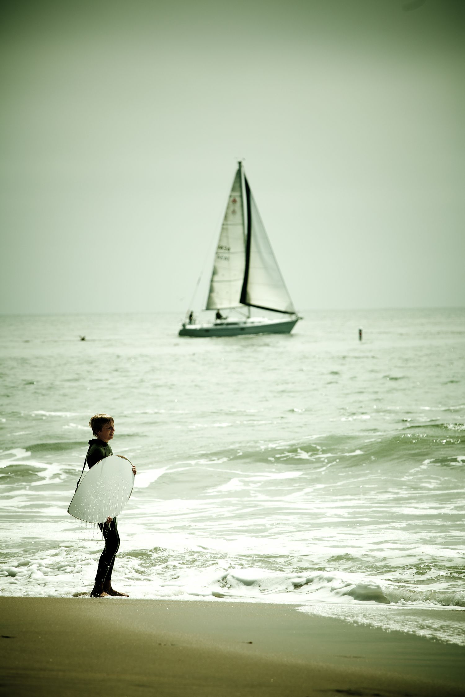 Young surfer with surfboard watching a sailboat on the Pacific Ocean in Santa Cruz, California along the scenic coastline.