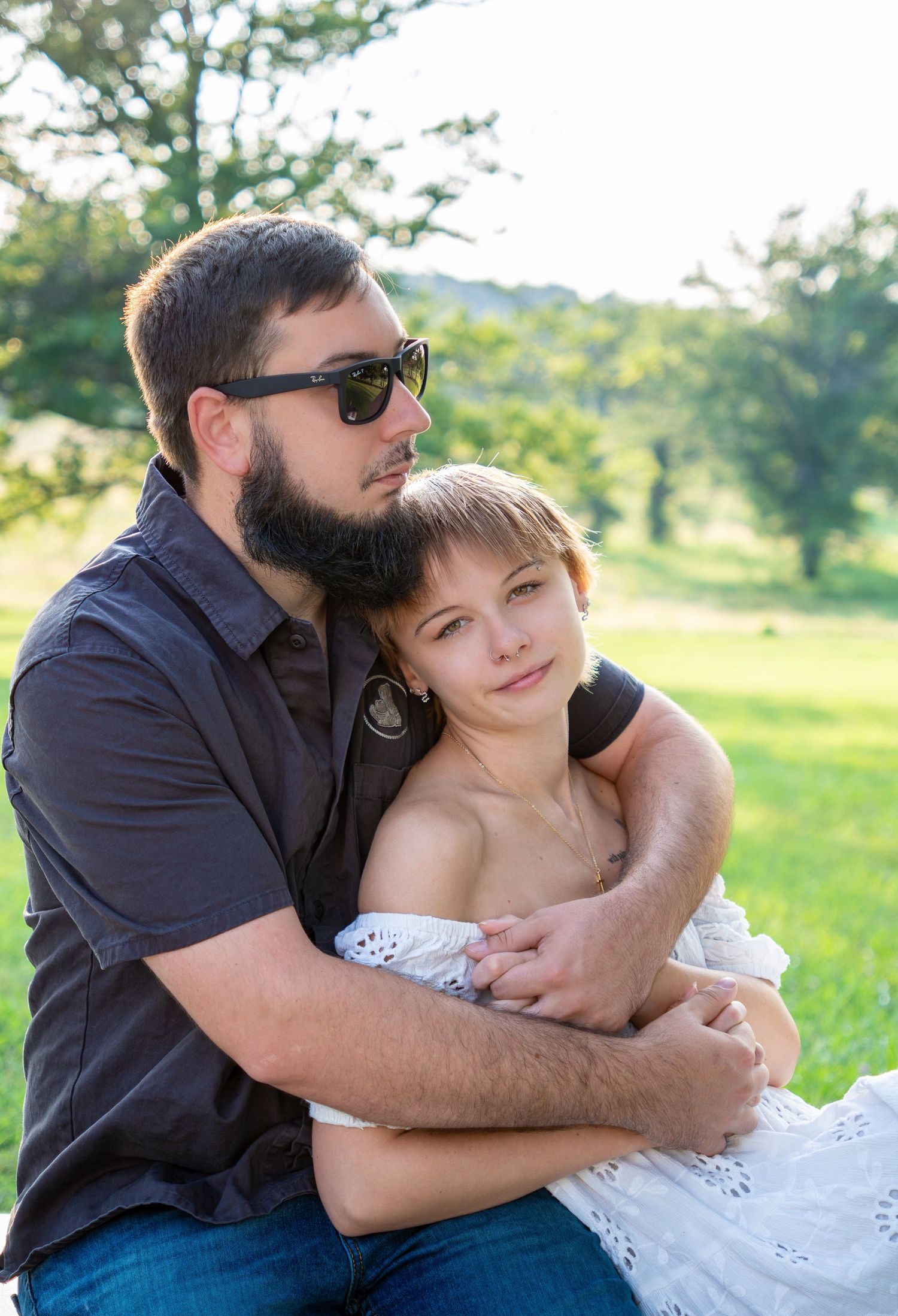 A bearded man in sunglasses tenderly embraces a woman in a white dress outdoors in a sunny green park.