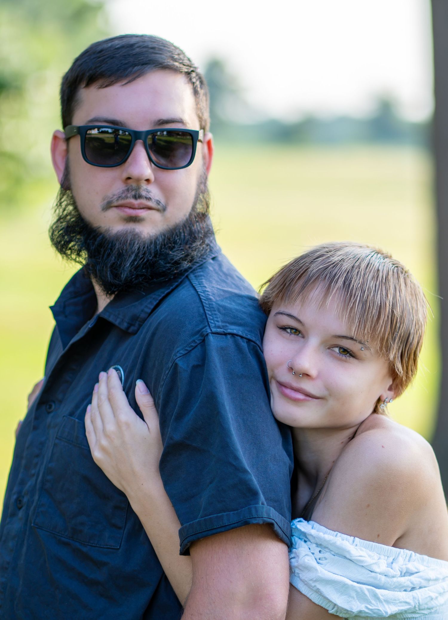 A bearded man in sunglasses and a woman with short hair embrace outdoors in a green field portrait.