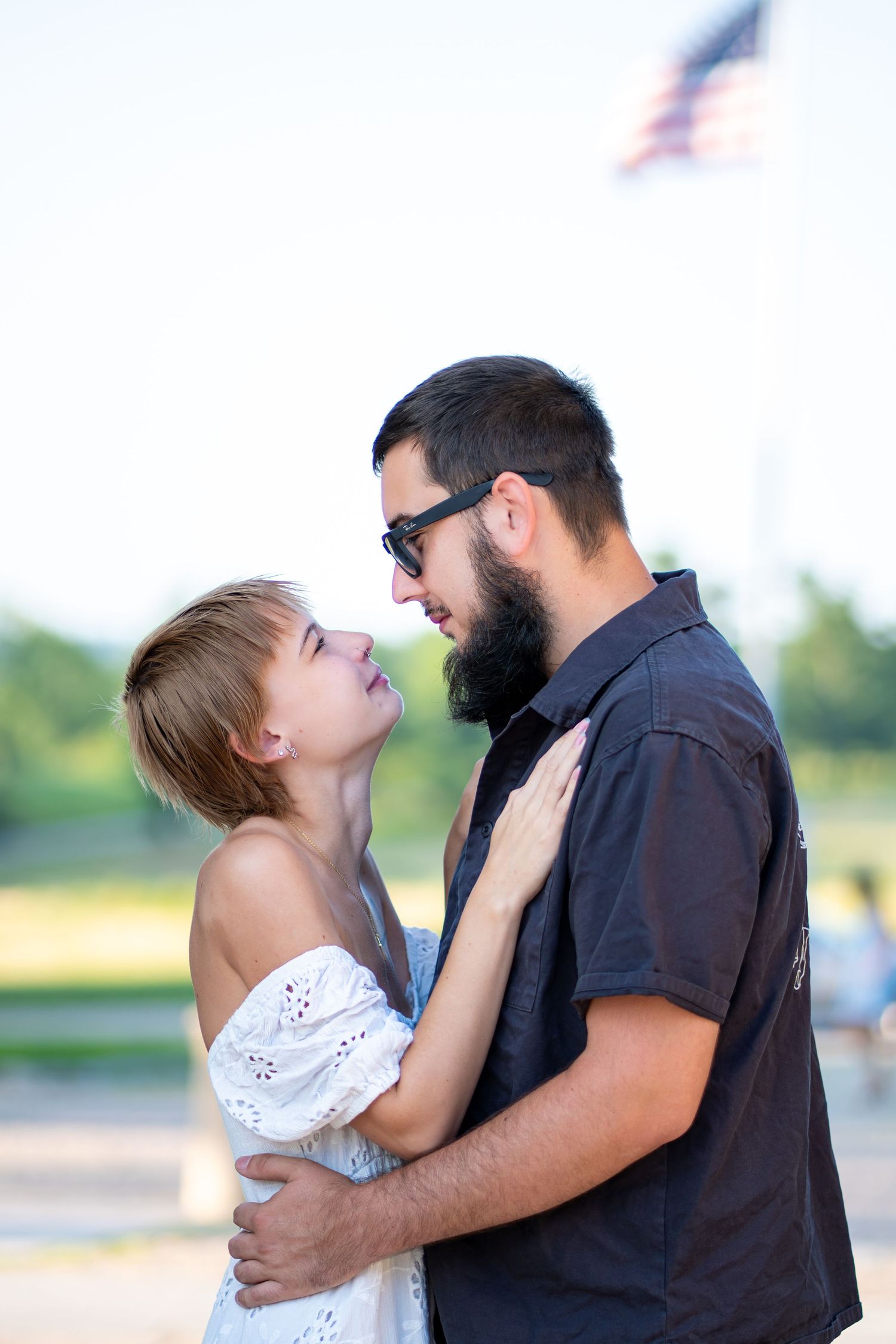 A couple embraces and gazes into each other's eyes outdoors with an American flag in the background.