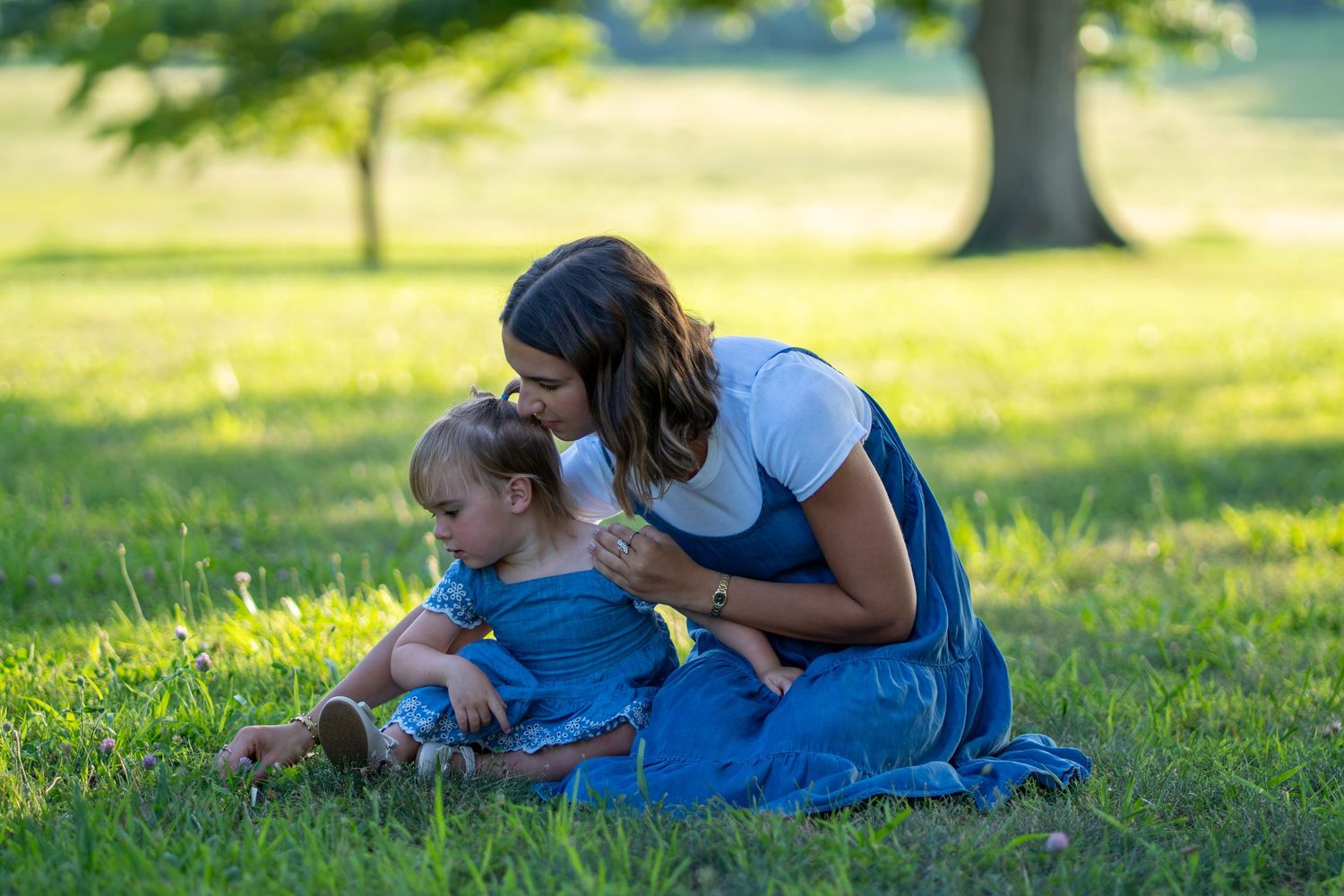 A mother sits on green grass in a park, tenderly kissing her baby girl dressed in denim.