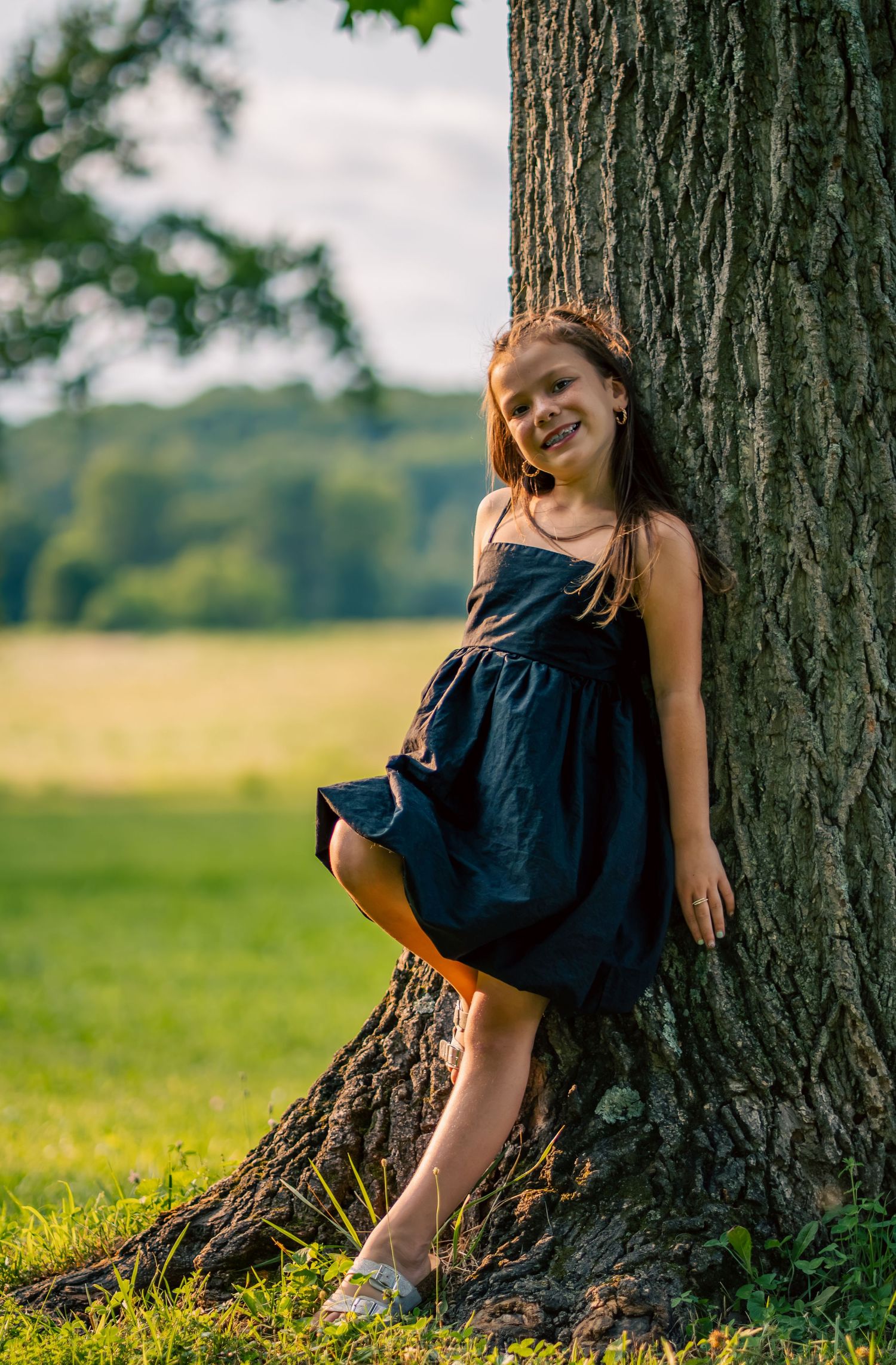 A smiling young girl in a navy dress leans against a large tree trunk in a sunny green park.