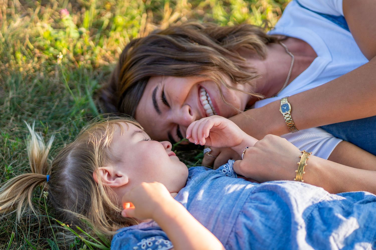 A mother and daughter laughing together while lying in green grass on a sunny day.