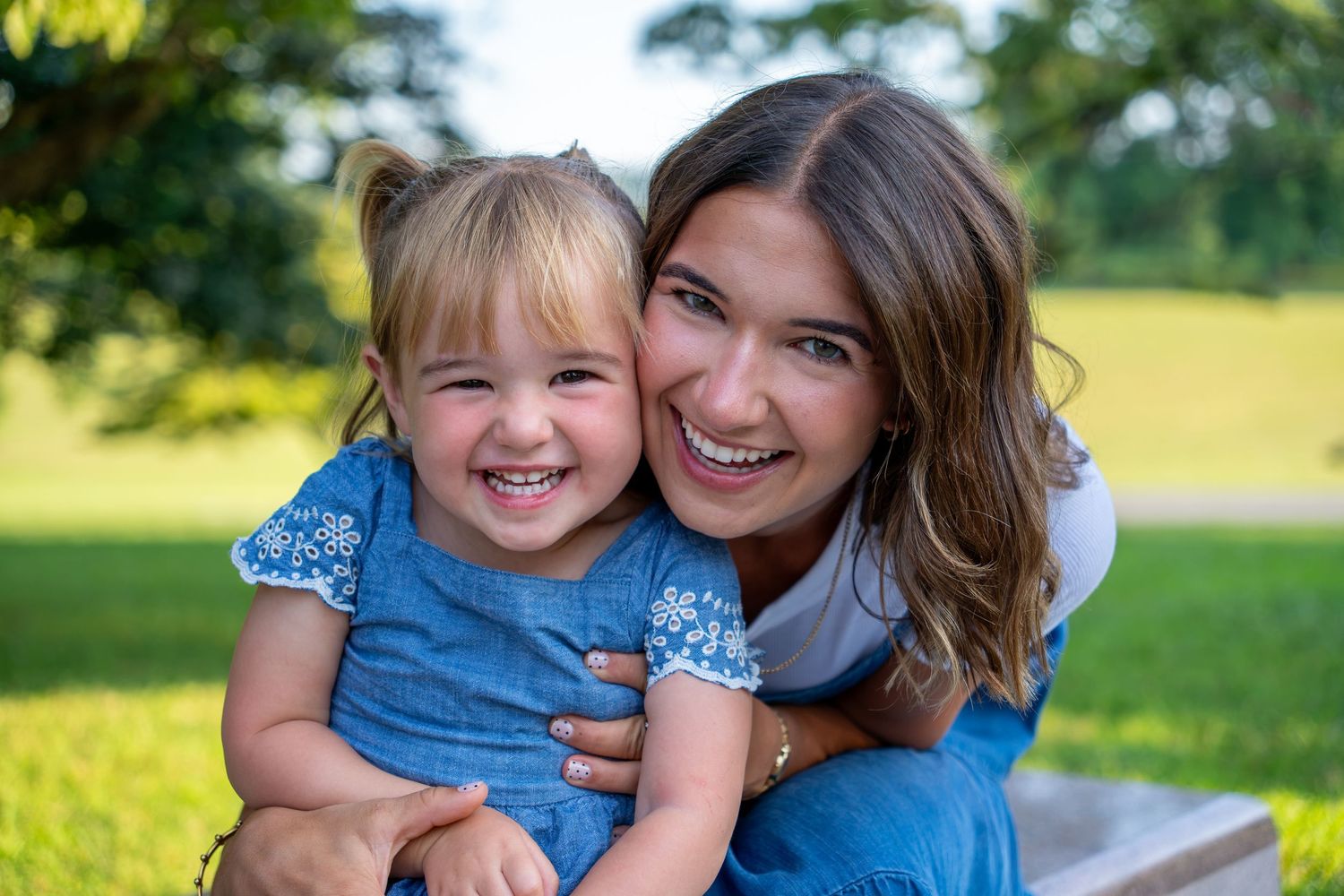 A young woman and toddler girl in matching blue dresses smile joyfully together outdoors in a green park.