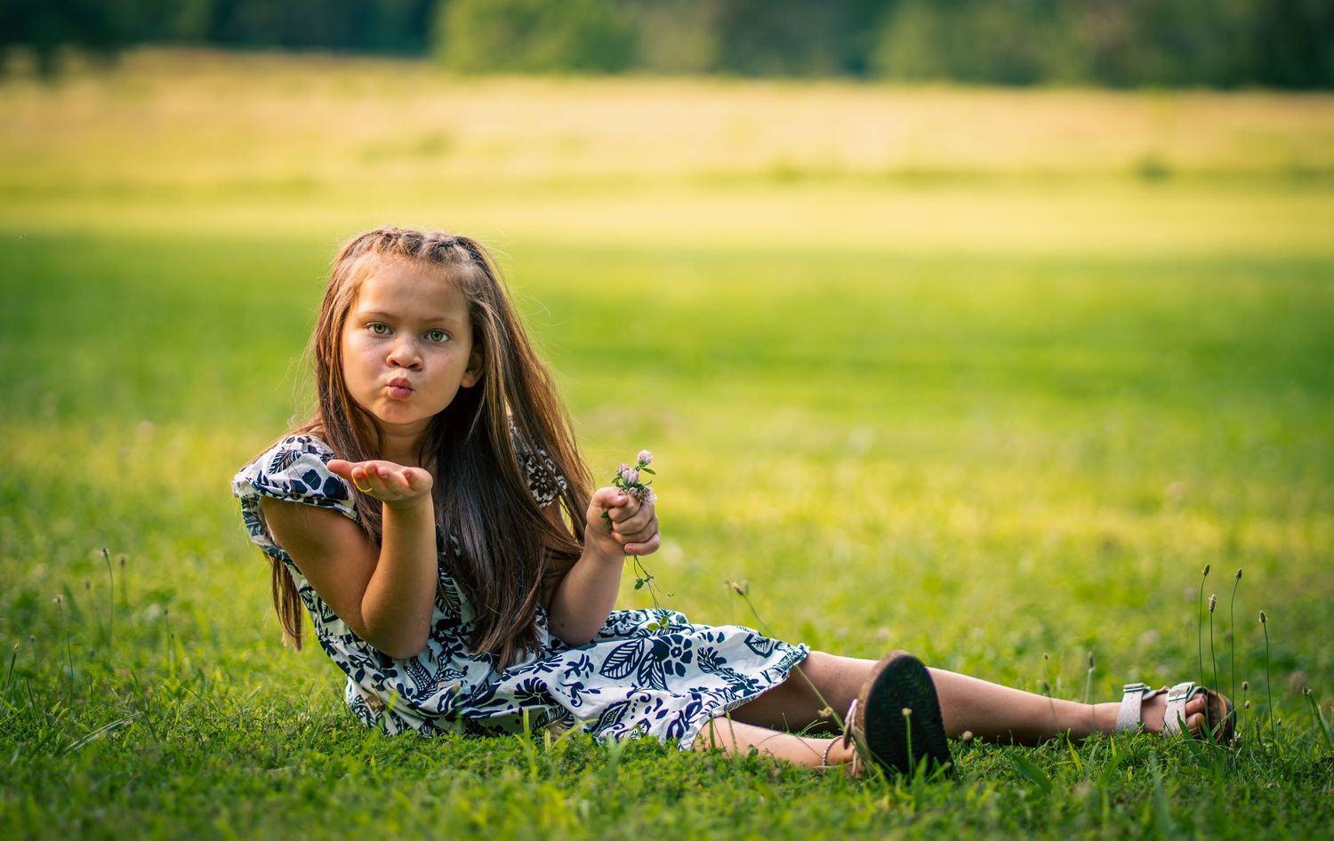 Young girl in floral dress sitting on green grass, blowing a kiss while holding a small flower outdoors.