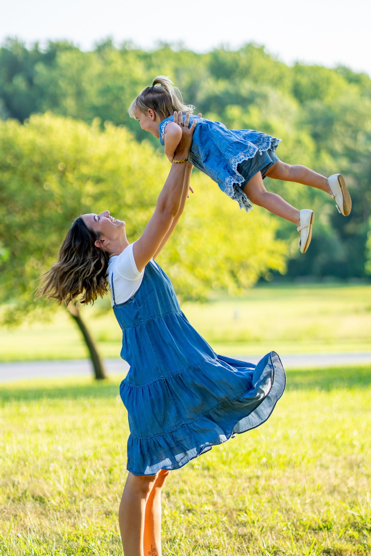 Mother in blue dress joyfully lifting her young daughter high in the air in a sunny green park.