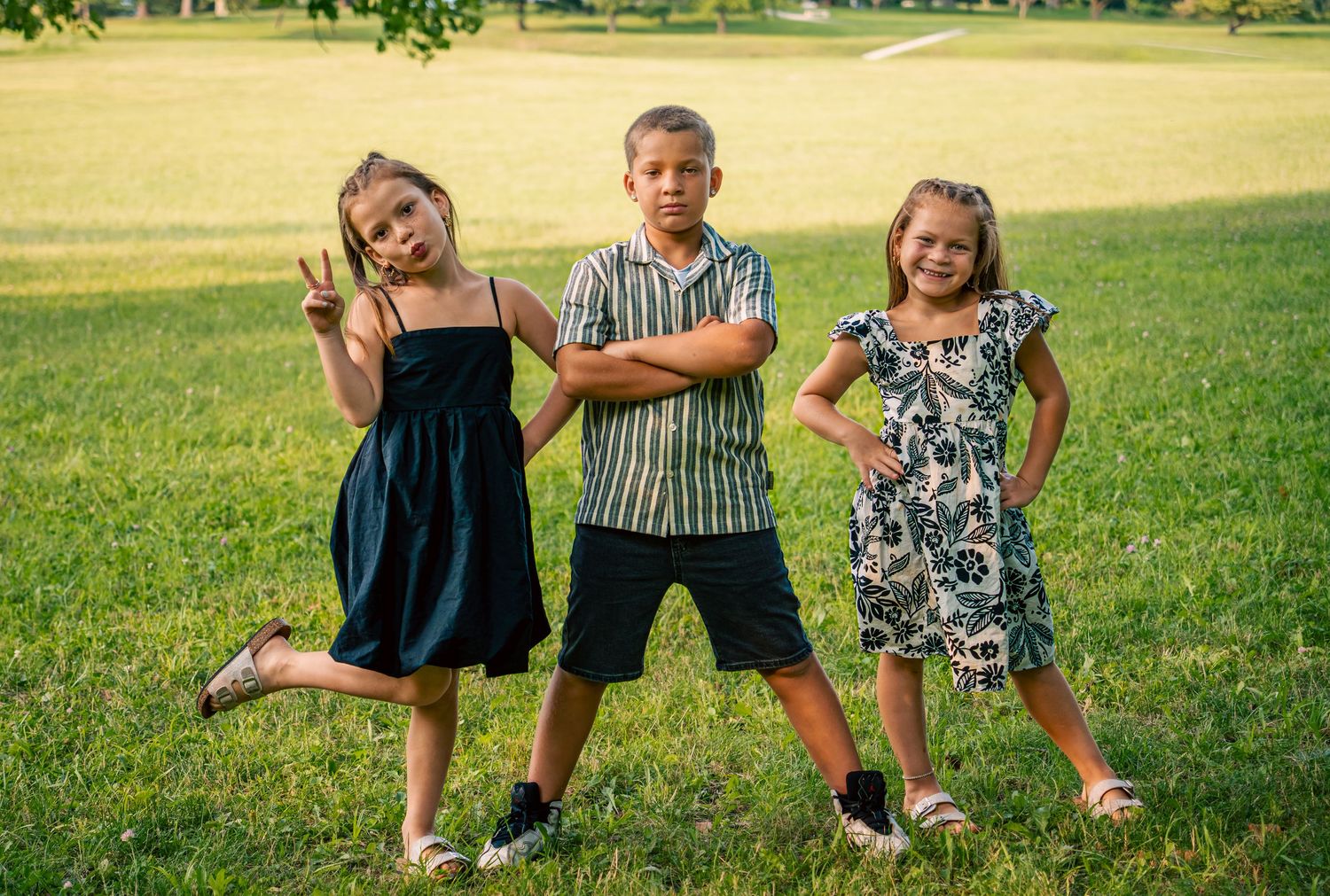 Three children posing outdoors on green grass, two girls in dresses and a boy with arms crossed in striped shirt.