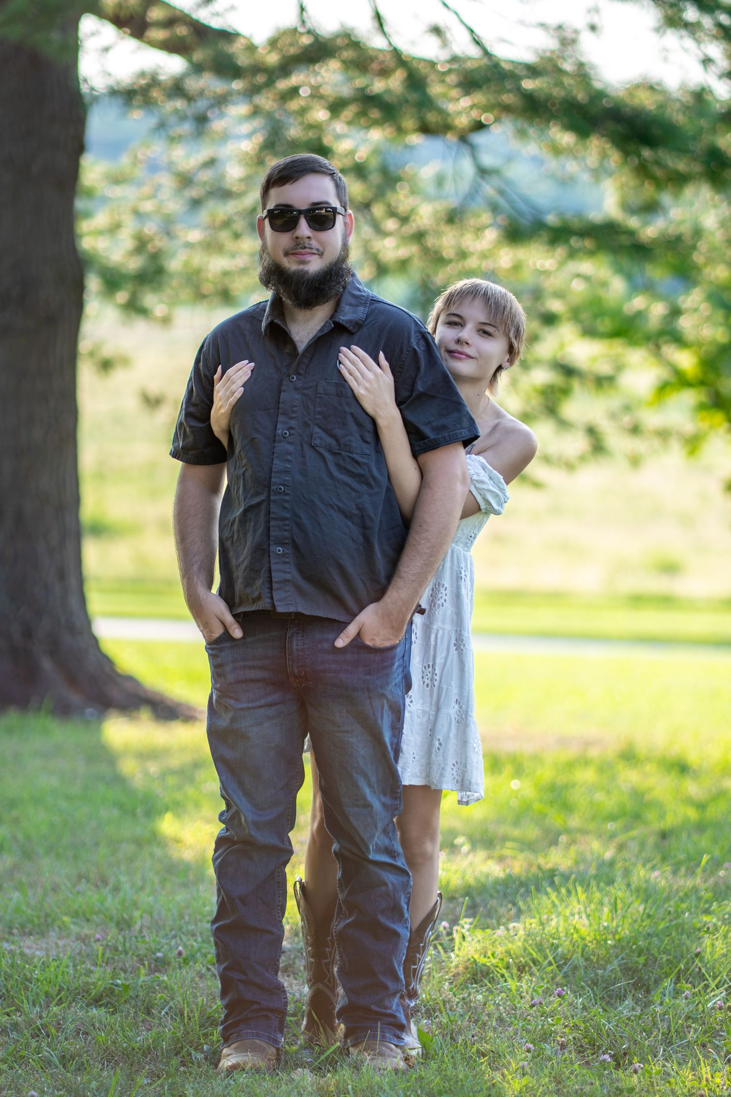 A bearded man in sunglasses and a woman in a white dress pose together outdoors under a large tree.