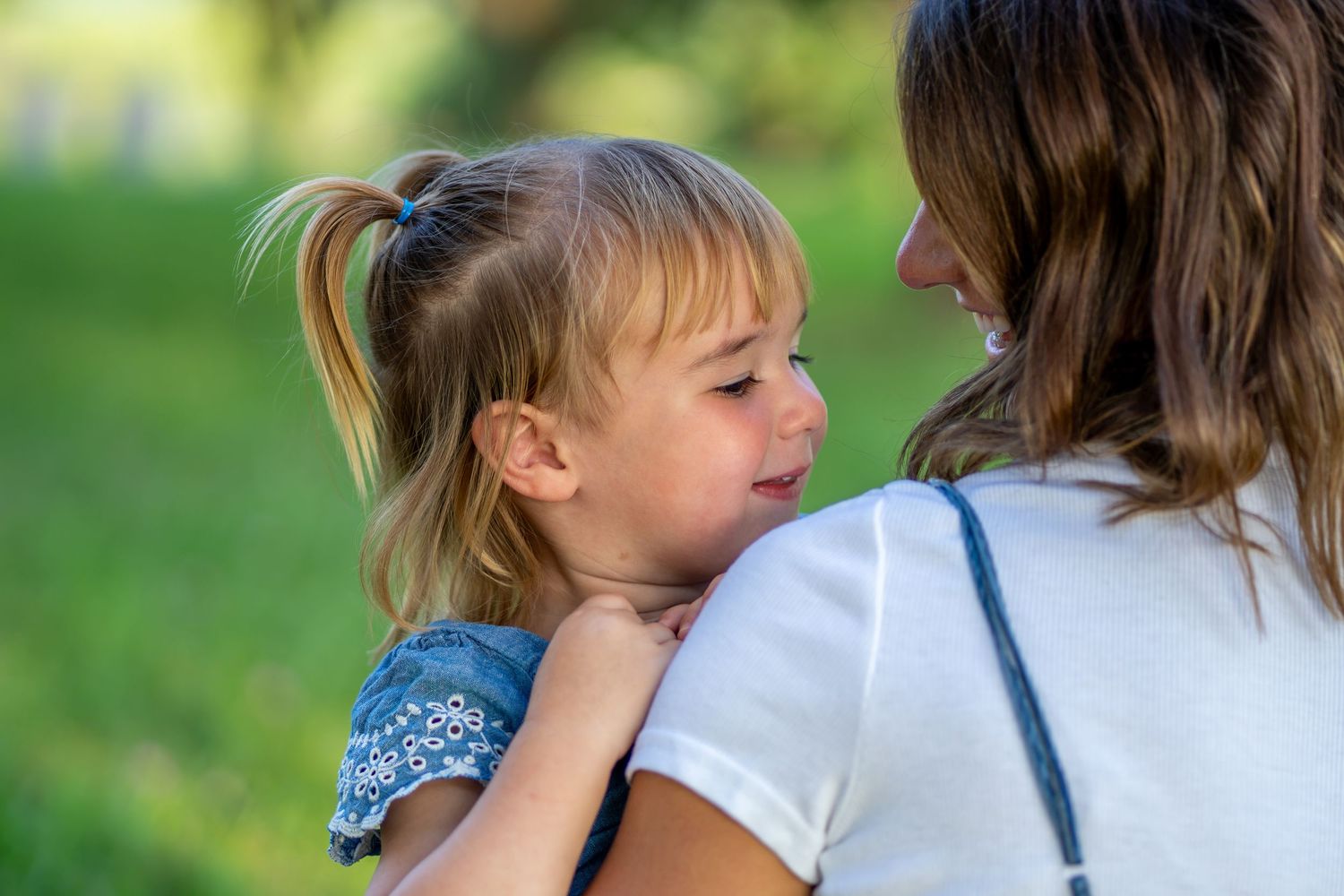 A mother holds her young blonde girl with pigtails in a sunny green park, sharing a tender moment.