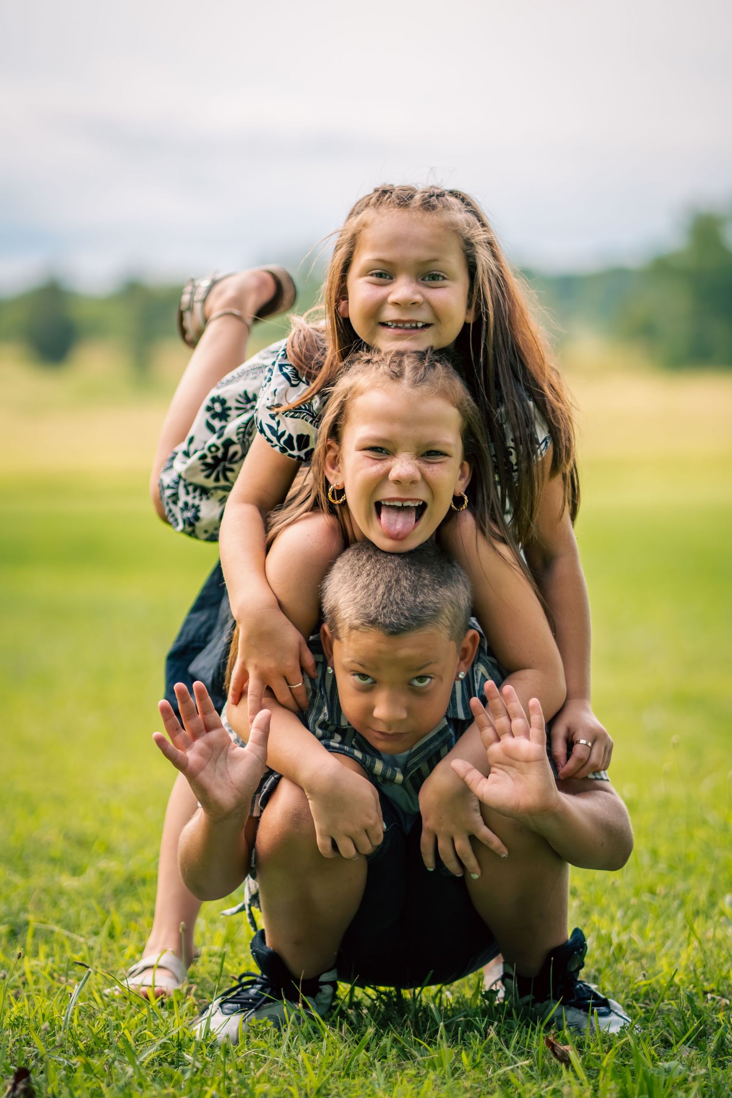 Three joyful kids stacking on each other in a grassy field, making funny faces and gesturing with their hands outdoors.