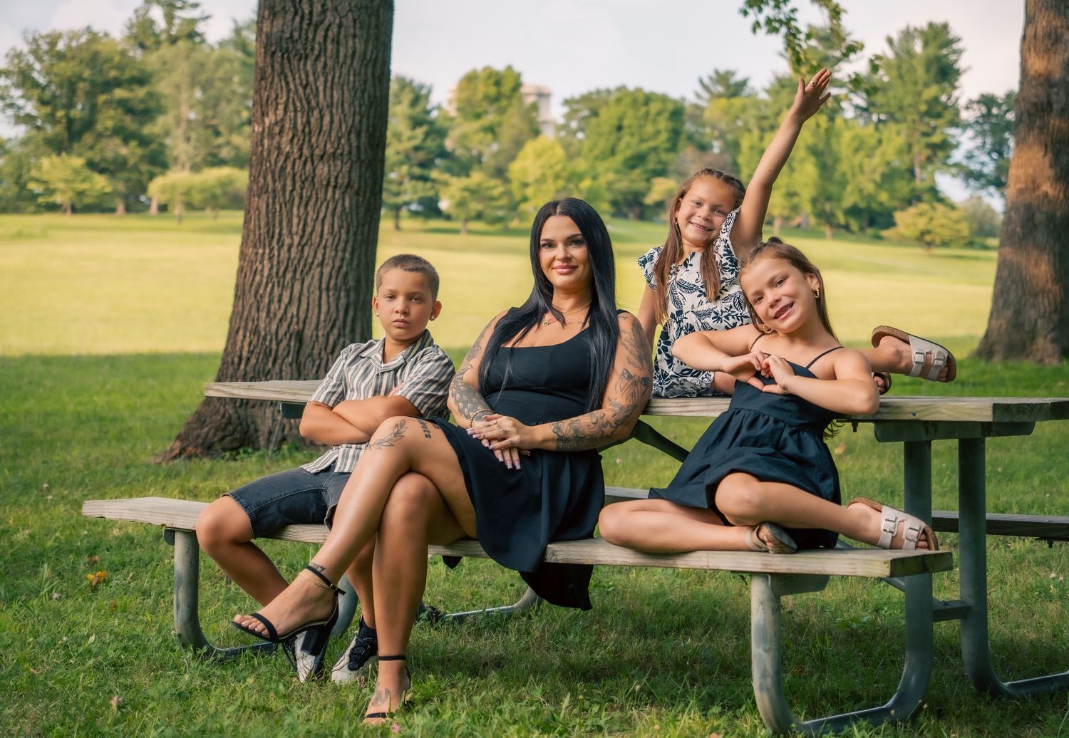 A woman and three children pose together at a picnic table in a lush green park on a sunny day.