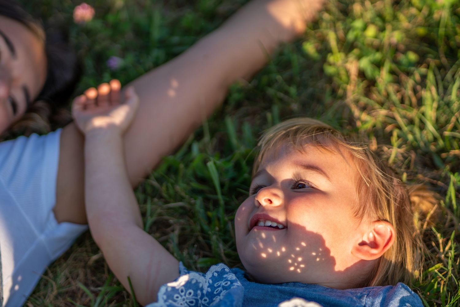 Two children lying on green grass in golden sunlight, with dappled light patterns on a smiling toddlers face.