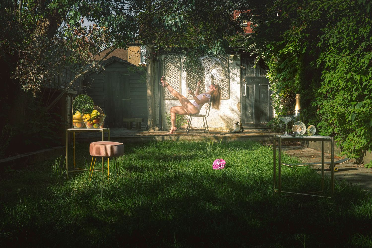 Woman reclining in garden chair pouring champagne, surrounded by luxury props on overgrown lawn, fine art conceptual photography London
