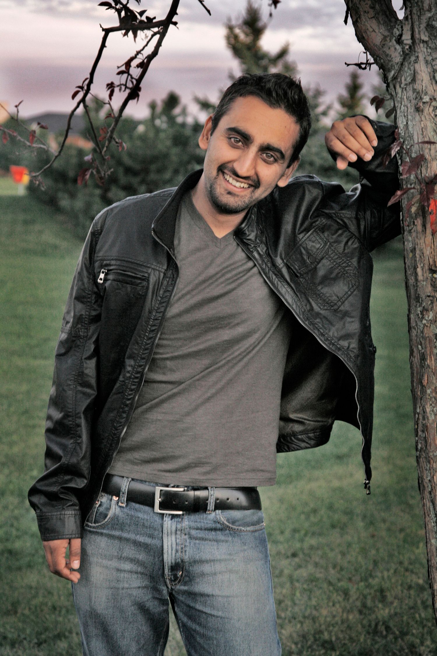 Color photo of a young man leaning on a tree with one arm, in a black leather jacket smiling widely.