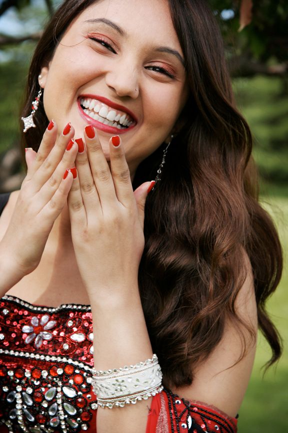 Photo of a young woman, wearing red and black bejeweled south asian ethnic wear, captured laughing with her hands in front of her.