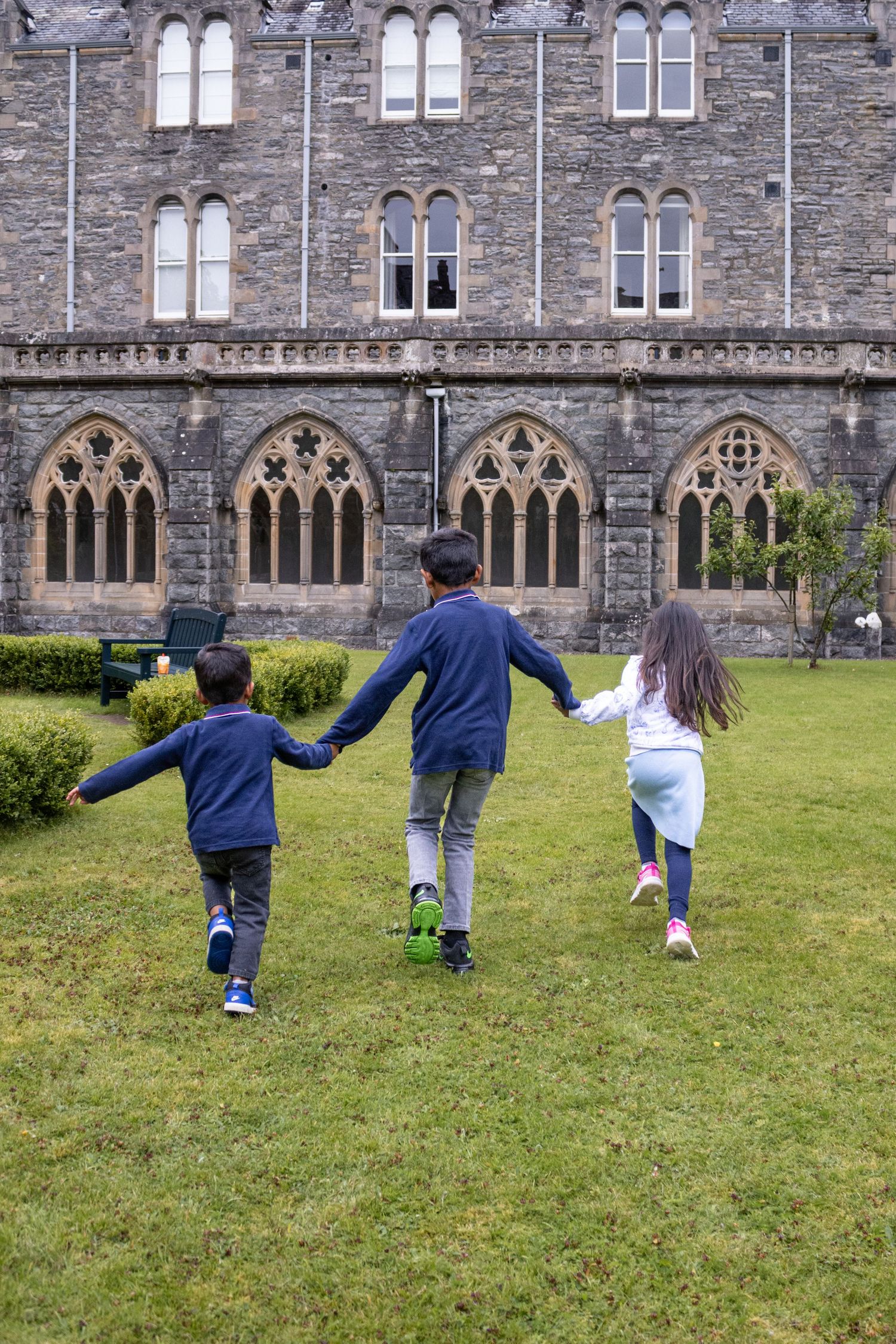 Three children holding hands run toward a grand Gothic stone building across a green lawn.