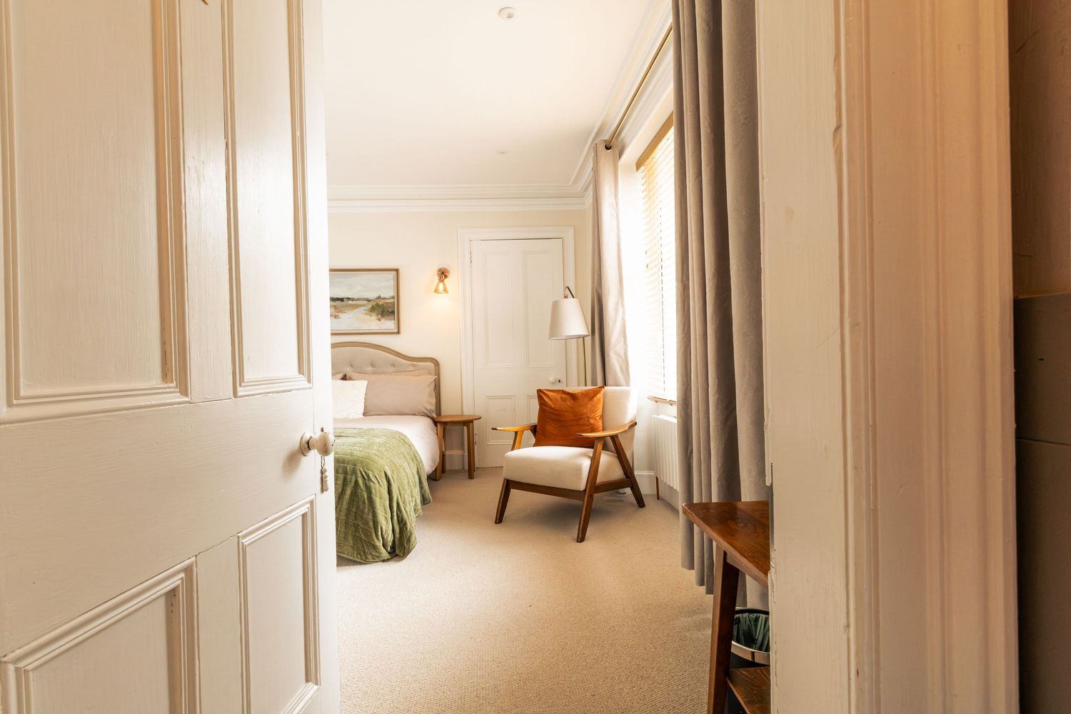 Elegant hotel suite with cream walls, plush armchair, green accented bed, and natural light from draped windows.