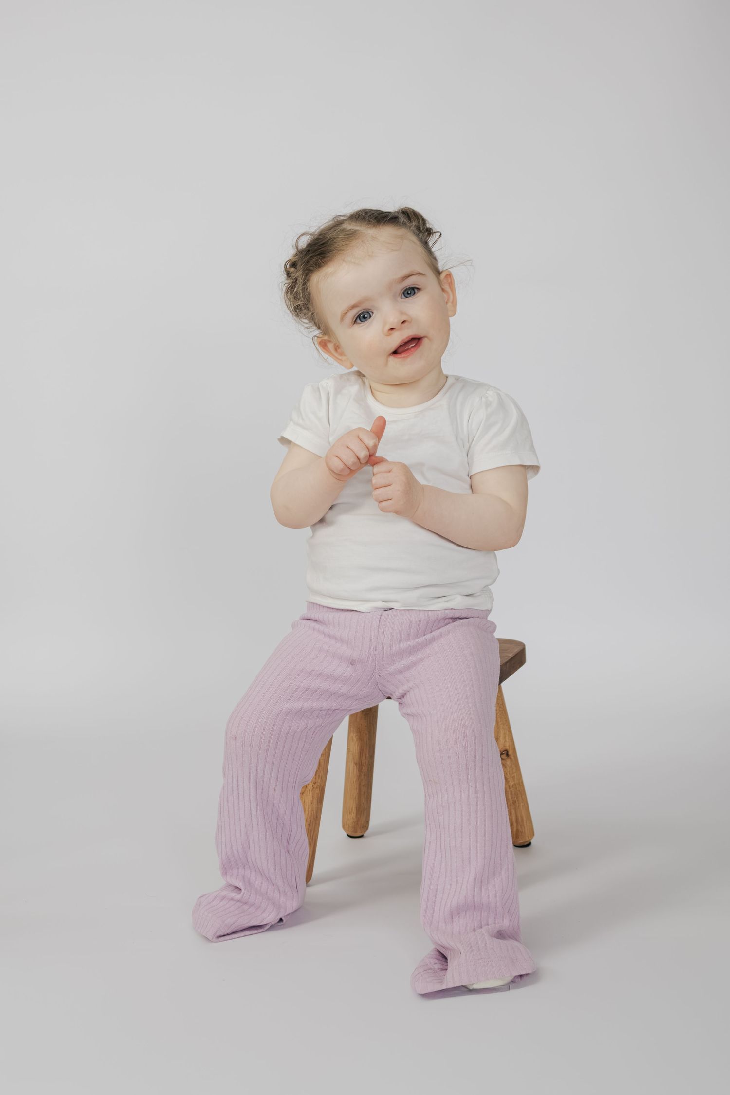 Cute baby girl in white tee and pink ribbed pants sitting on wooden stool giving thumbs up.