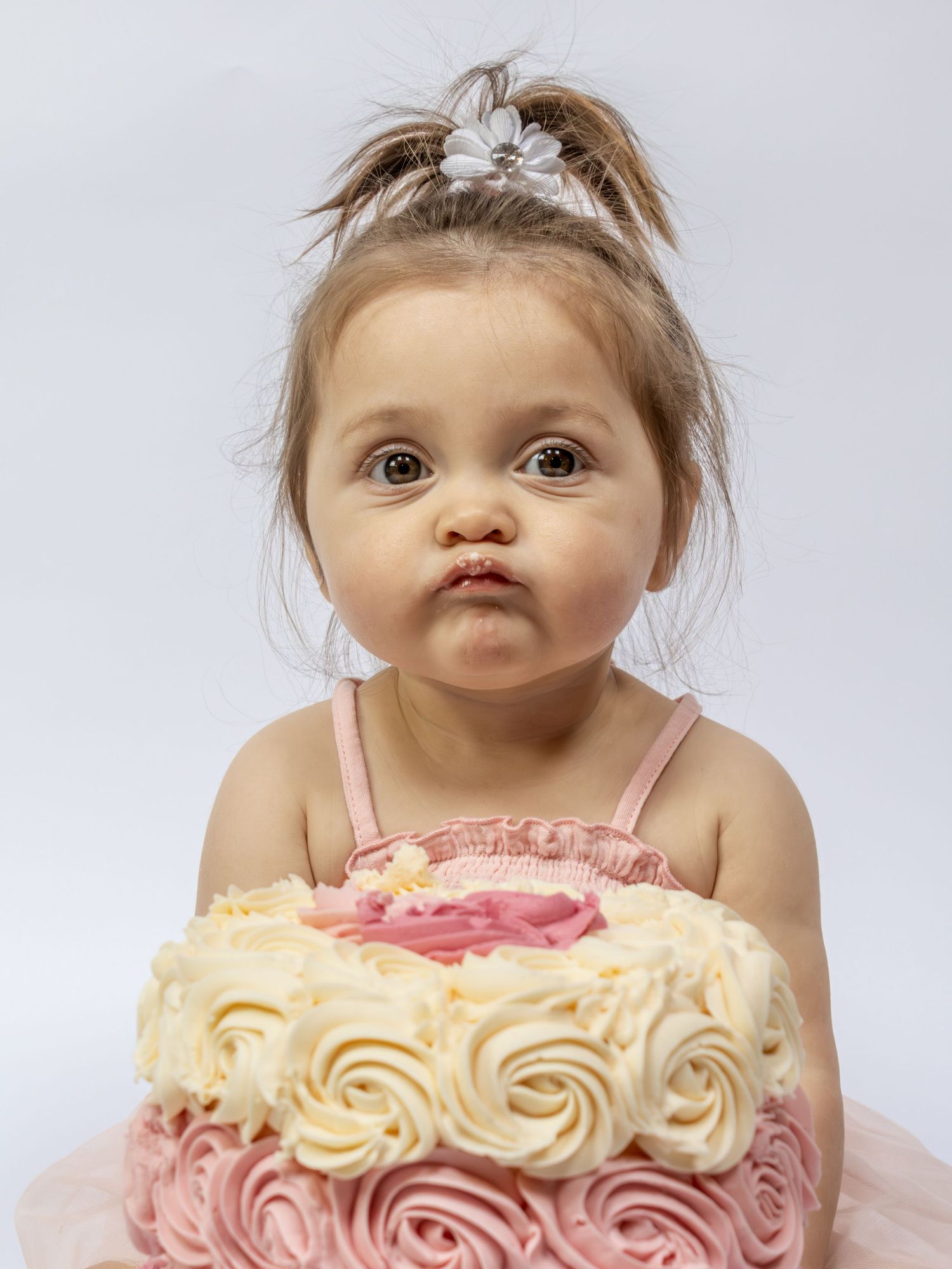 Adorable toddler girl in pink dress and tiara making a pouty face in front of a pink rosette birthday cake.