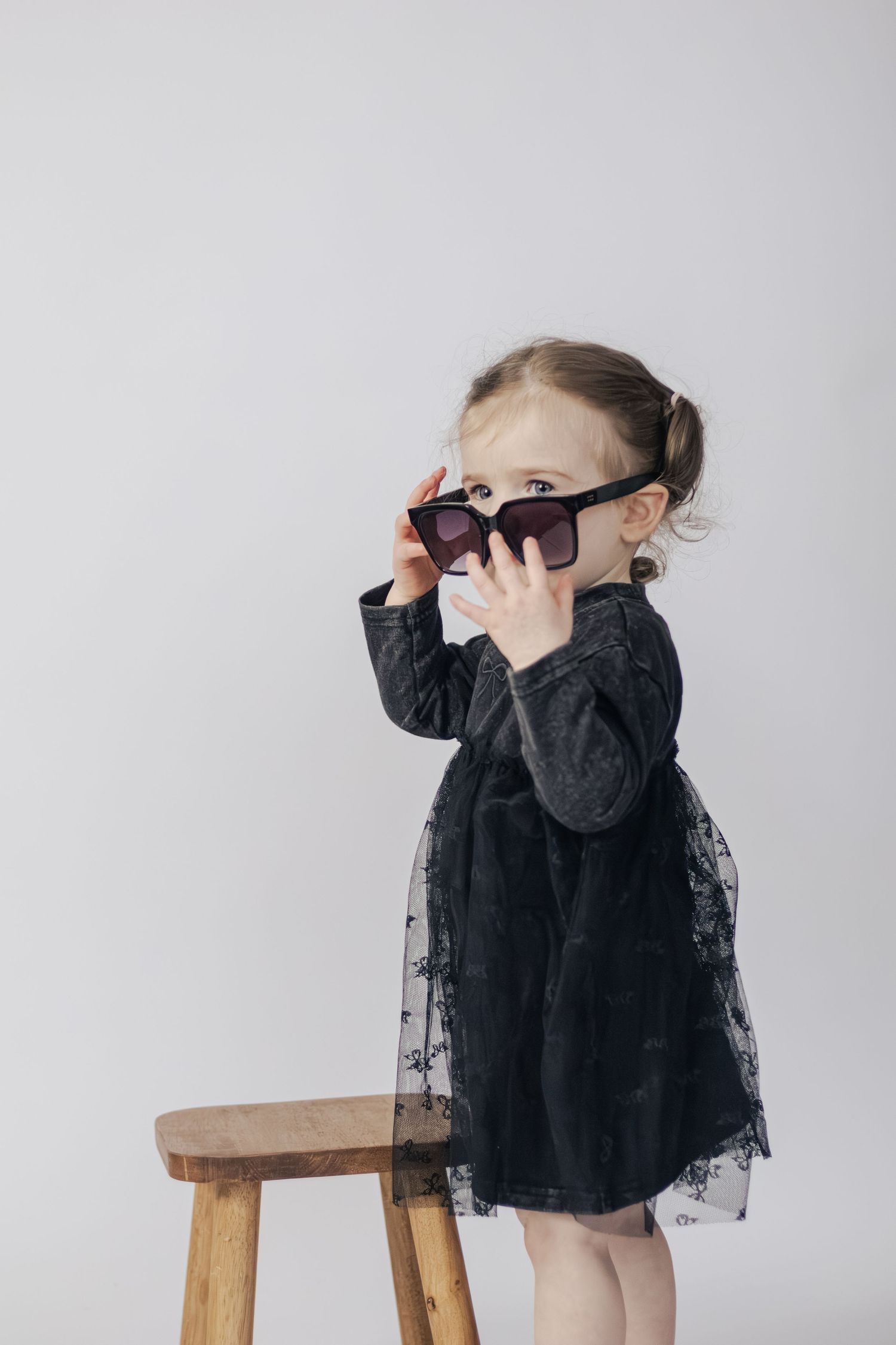 Toddler girl in black lace dress peering over oversized dark sunglasses beside a wooden stool.