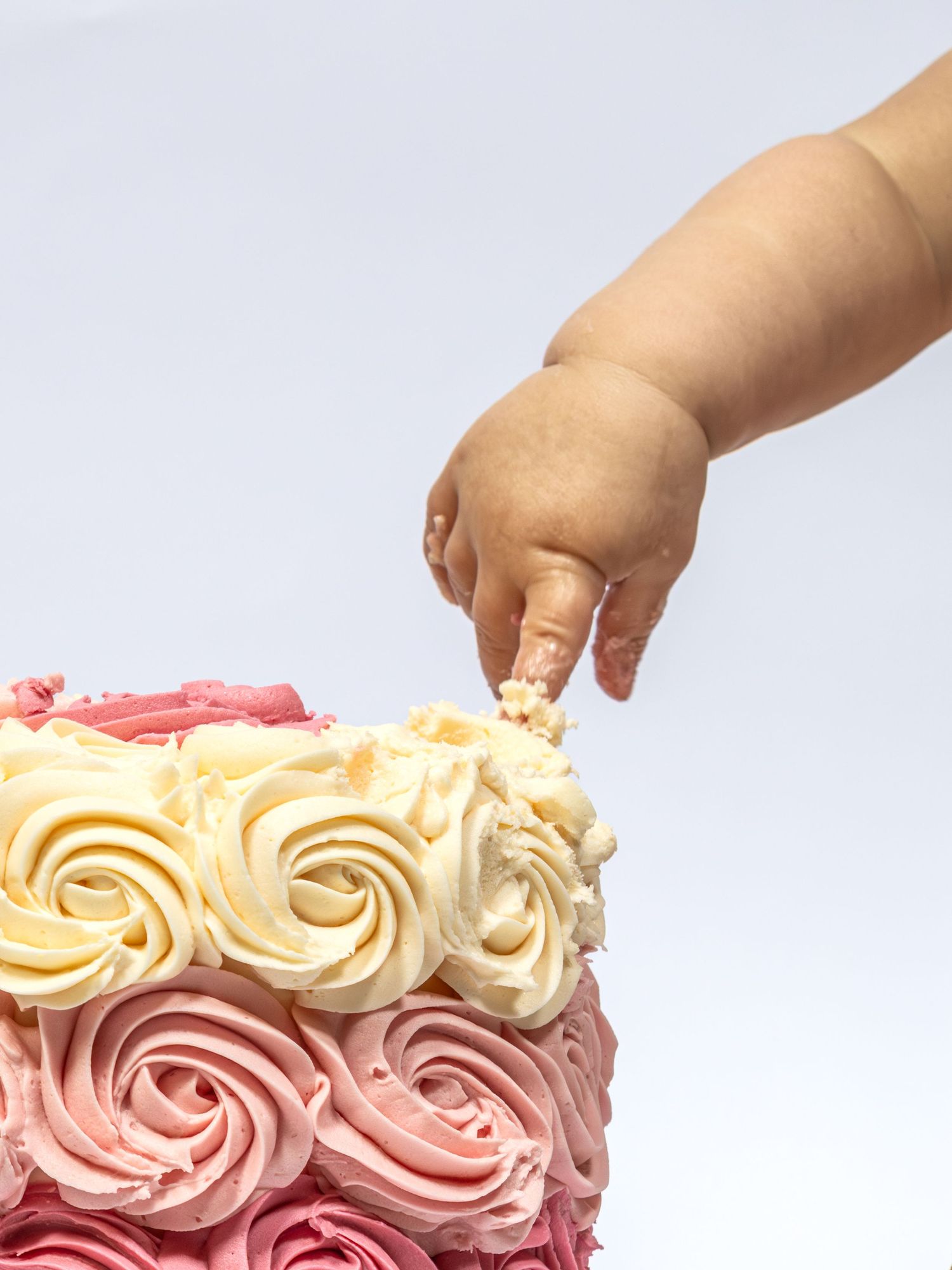 A baby's hand reaching into a pink and white rose swirl frosted birthday cake on a white background.