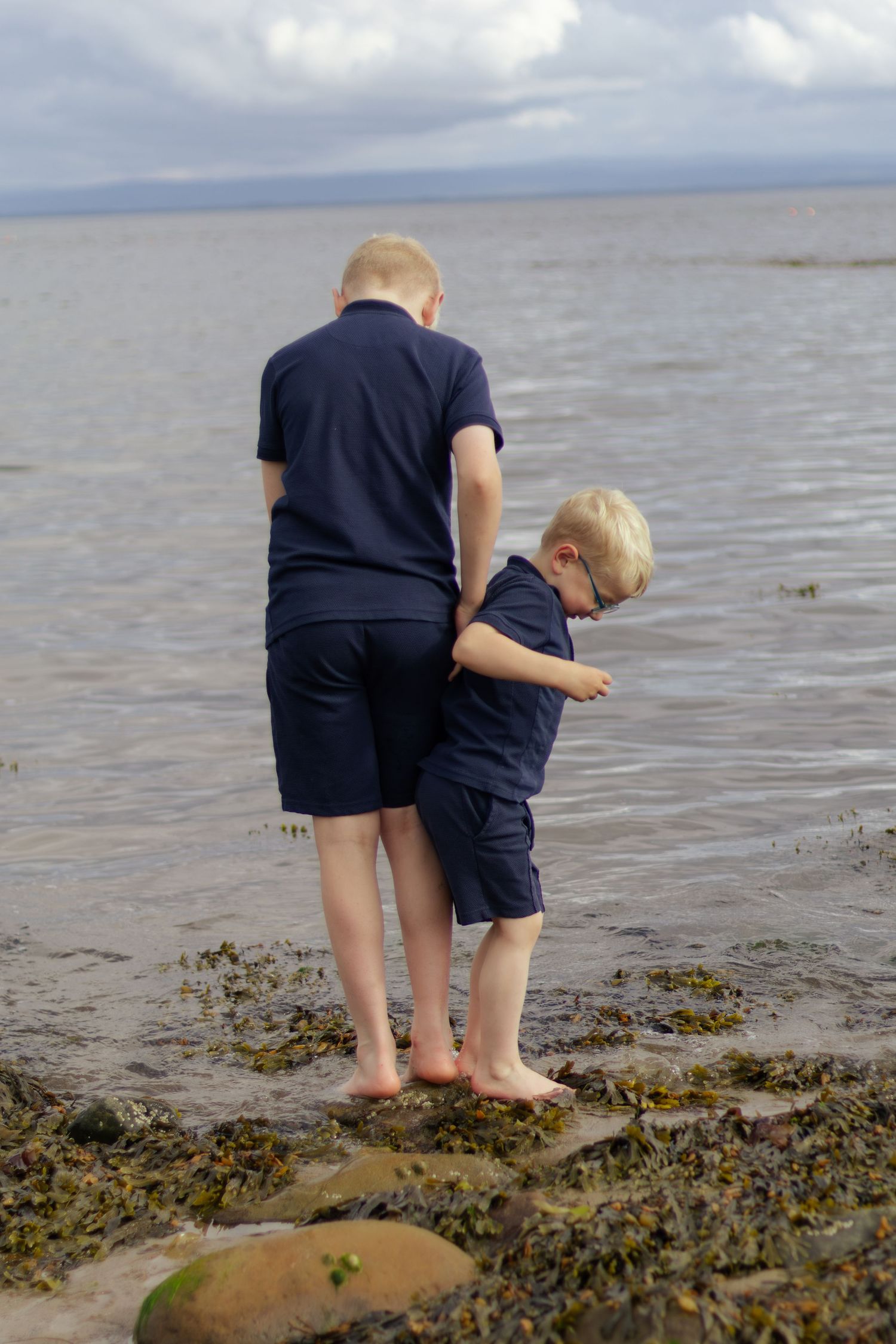 Two blonde boys in matching navy outfits explore rocky shoreline barefoot, paddling in shallow coastal waters on a cloudy day.
