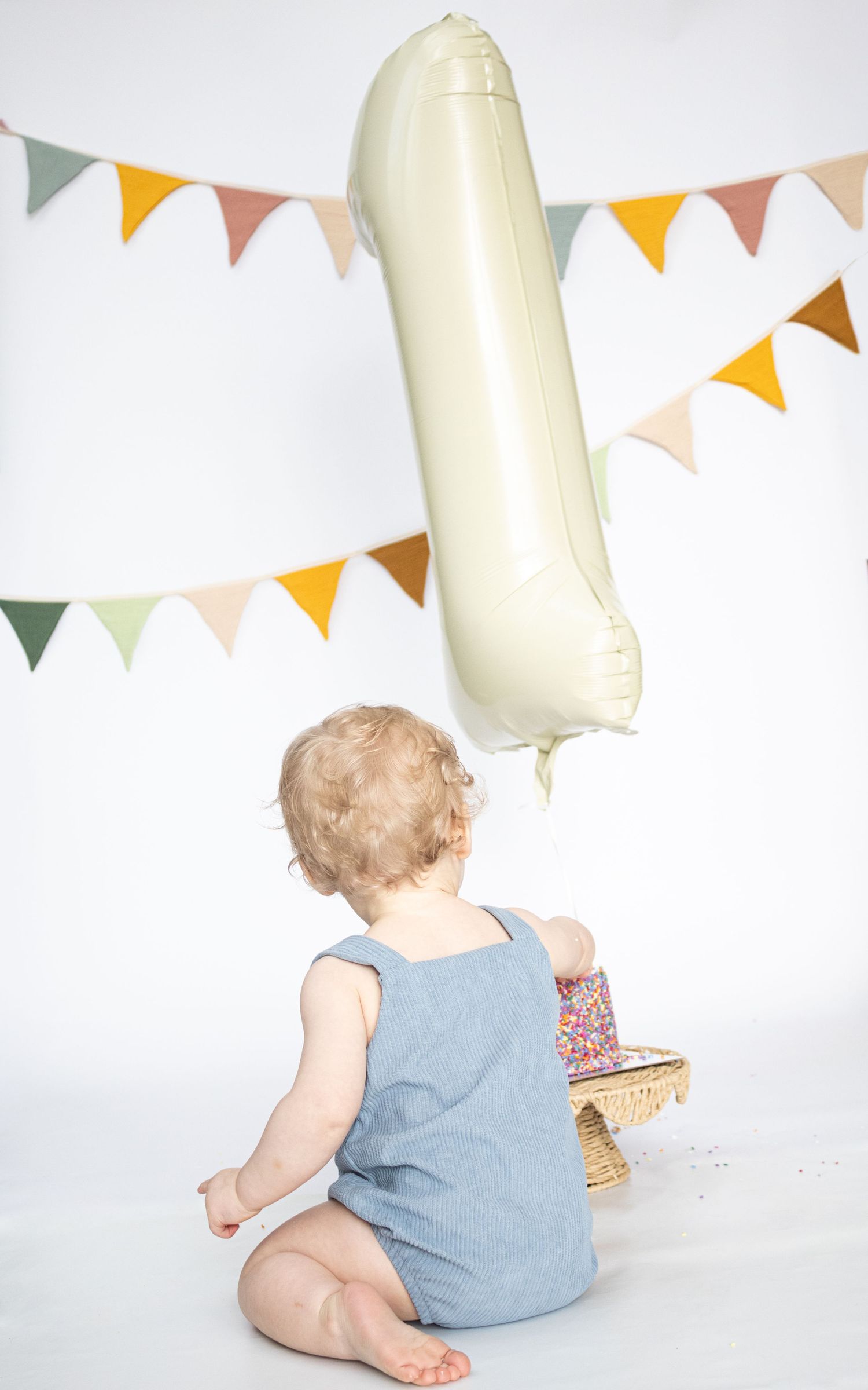 Baby in blue romper reaches for number 1 balloon at first birthday party with colorful bunting.