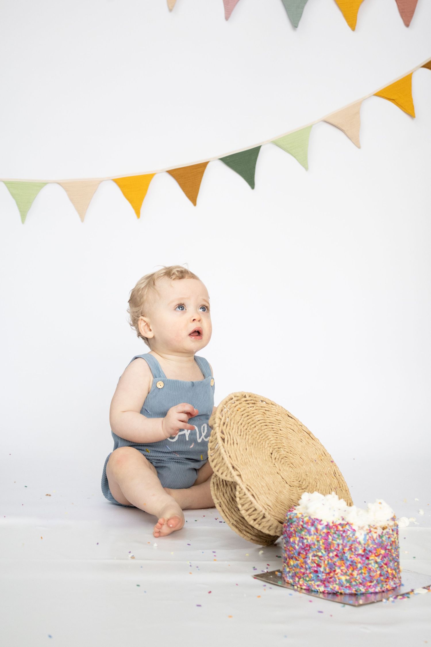 Curious one-year-old baby in blue romper sitting beside smashed birthday cake with colorful sprinkles and bunting decorations.