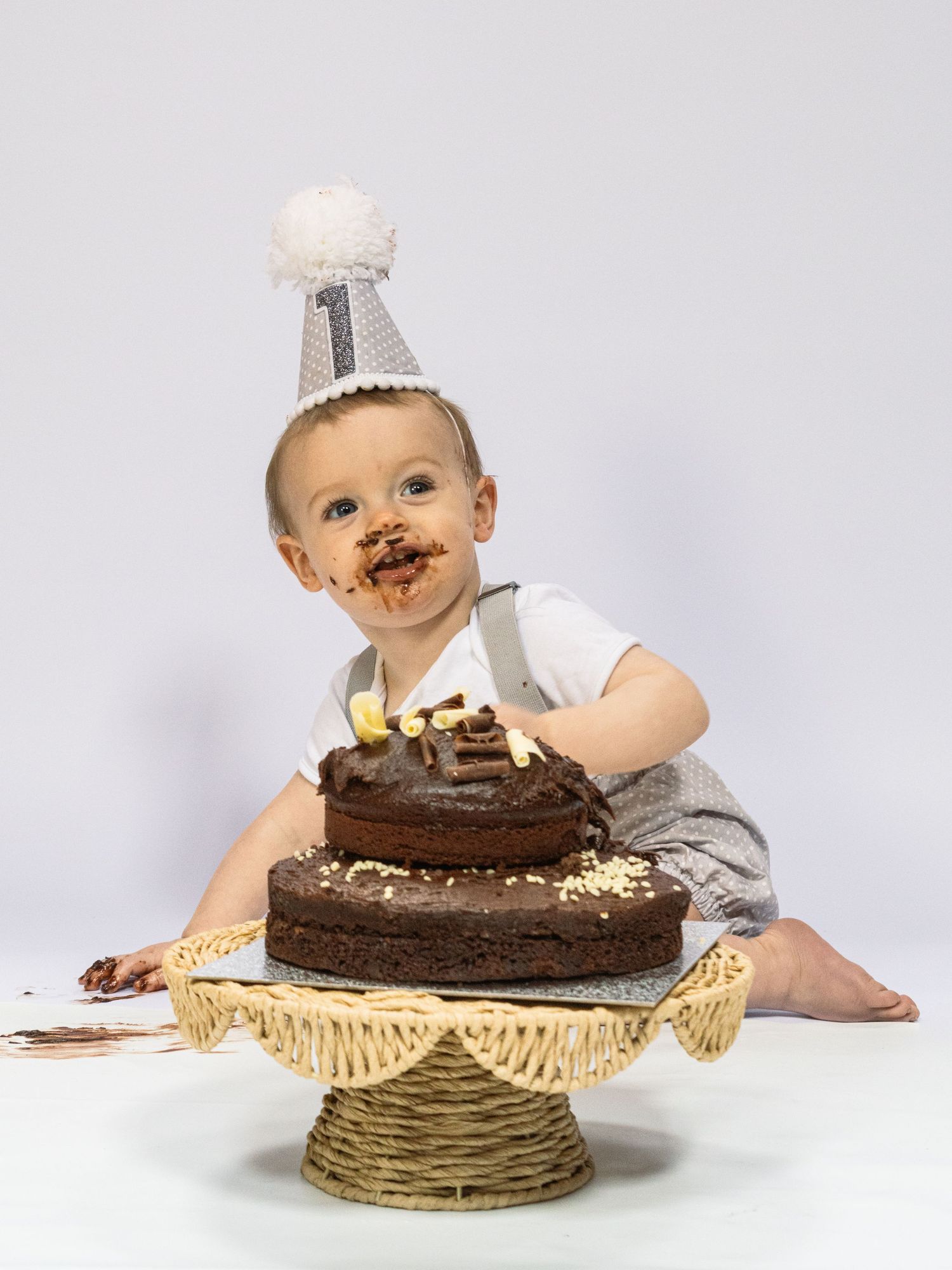 Baby celebrating first birthday smashing chocolate cake wearing a party hat with frosting on face.