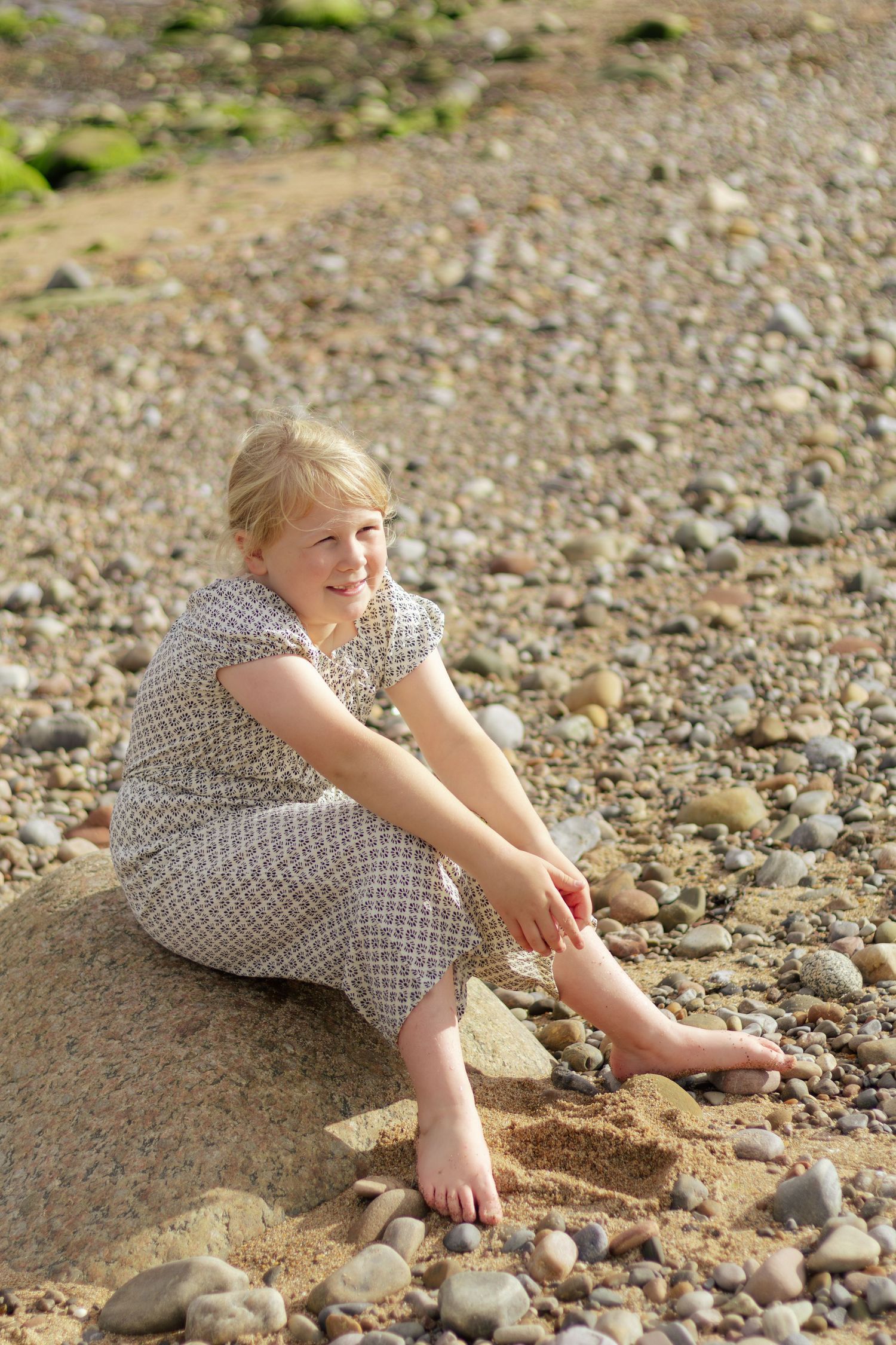 Young blonde girl in patterned dress sitting barefoot on a pebble beach, smiling outdoors.