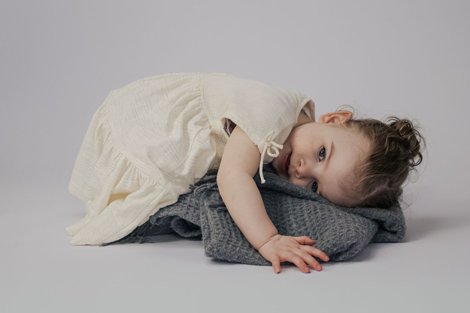 Toddler in cream dress resting head on grey knit blanket on floor.