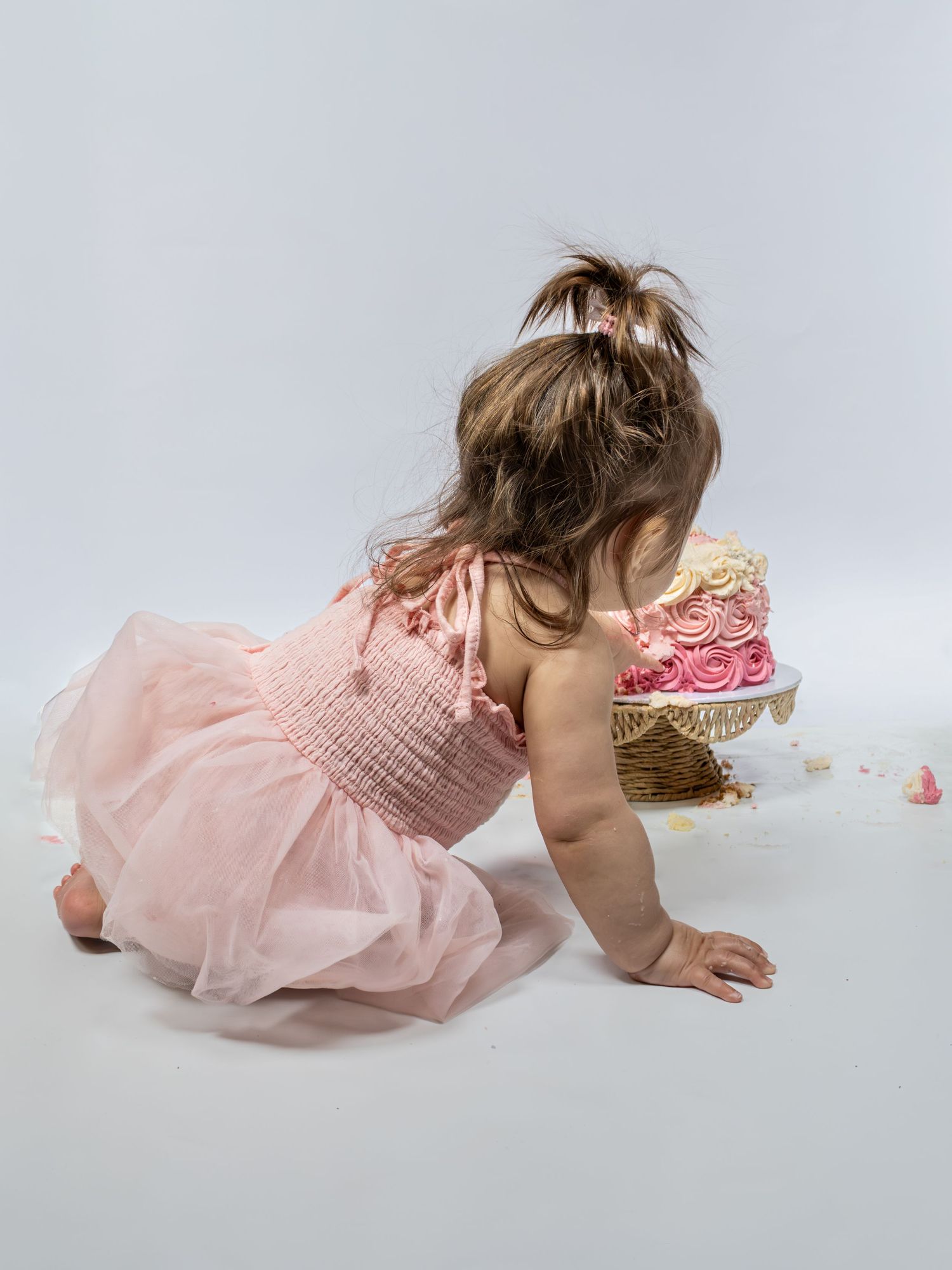 Baby girl in pink tutu dress reaching toward a decorated birthday smash cake with pink rosette frosting.