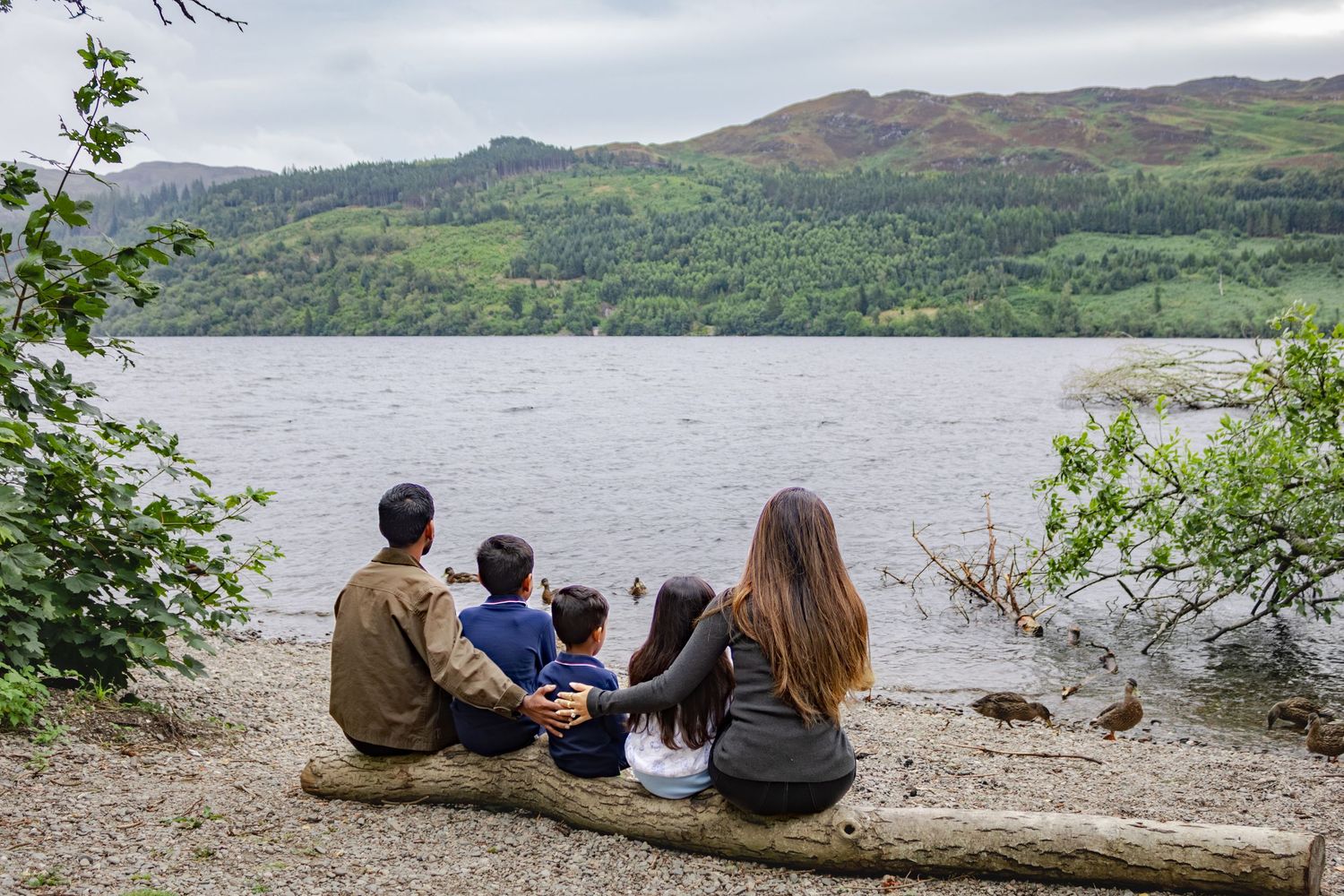 Family of four sitting on a log by a scenic loch, admiring green hills and calm water in the Scottish Highlands.